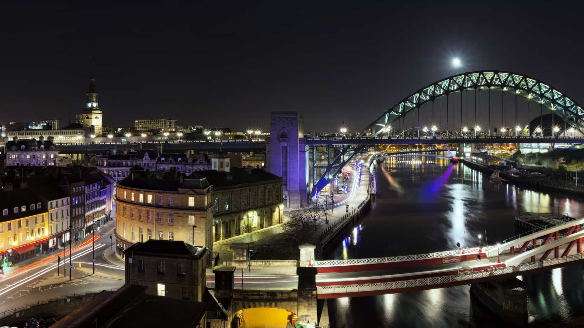 A beautiful nighttime view of the city of Newcastle Upon Tyne, England, with the Tyne Bridge and Swing Bridge illuminated, reflecting in the river under a full moon.