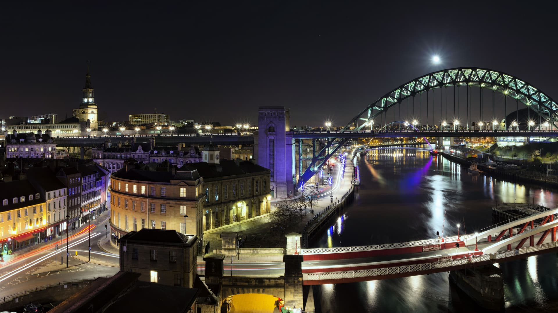 A beautiful nighttime view of the city of Newcastle Upon Tyne, England, with the Tyne Bridge and Swing Bridge illuminated, reflecting in the river under a full moon.