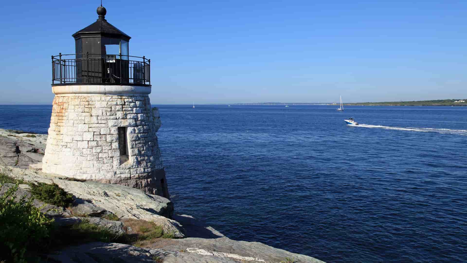 A scenic view of the Castle Hill Lighthouse in Newport, Rhode Island, with a white and black stone base, overlooking the deep blue Atlantic Ocean under a clear sky.