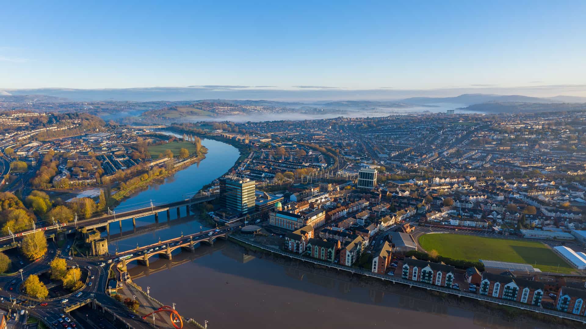 "An aerial panoramic shot of the city of Newport, Wales, with the River Usk winding through the urban landscape and a bridge connecting both sides of the city. "