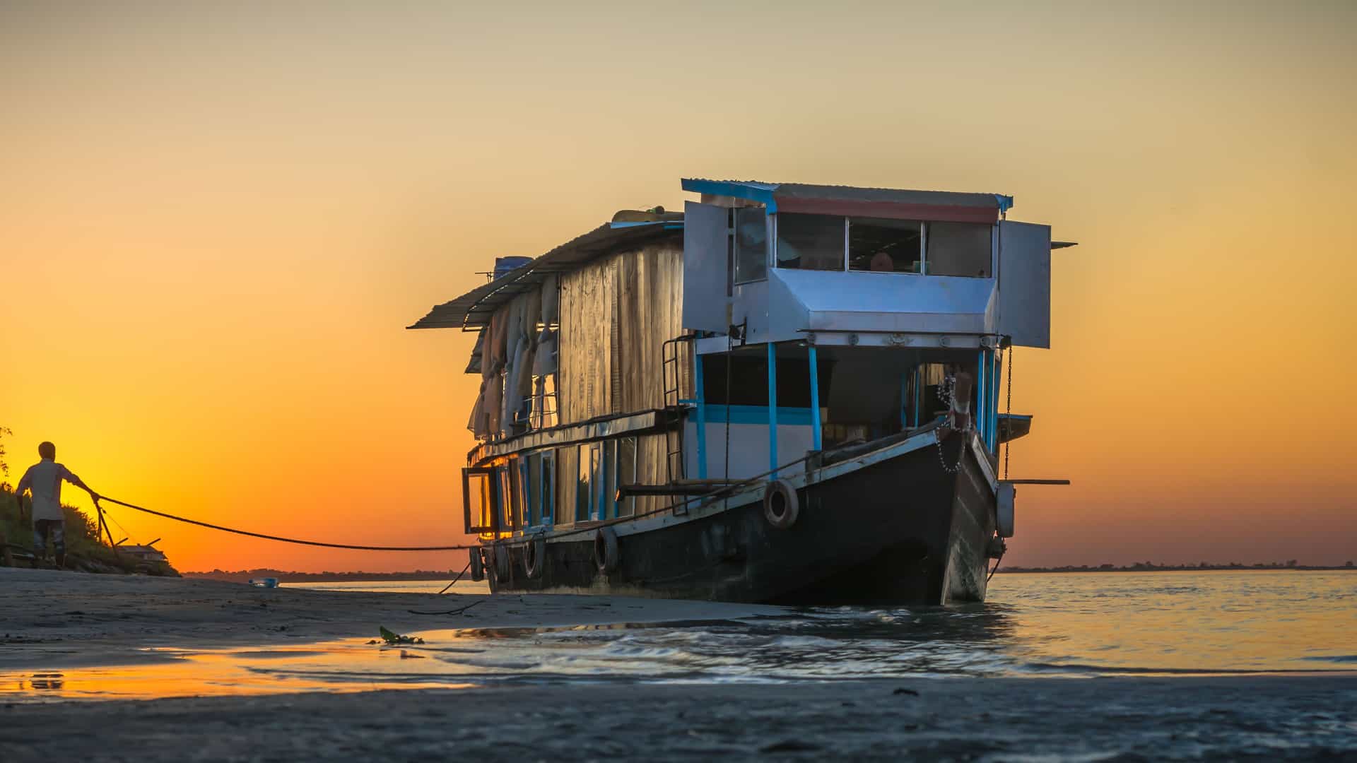 "A low-angle shot of a large ferry boat docked on the shore of the Brahmaputra River at Nimati Ghat, with a person pulling a rope against a beautiful sunset. "