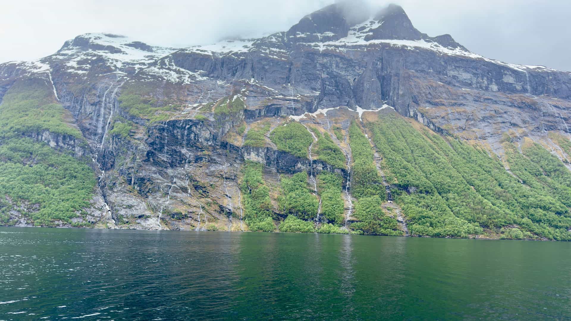 A large cruise ship navigates the calm waters of the Nordfjord in Norway, with a breathtaking view of the steep, rocky cliffs and a large waterfall cascading down into the fjord below.