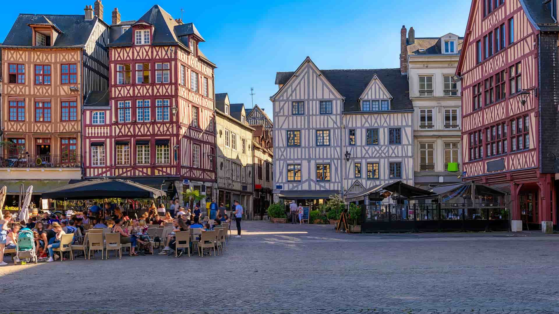 "A bustling summer day in Rouen, Normandy, with people dining at an outdoor cafe in front of historic, half-timbered houses in the Place du Vieux Marché. "