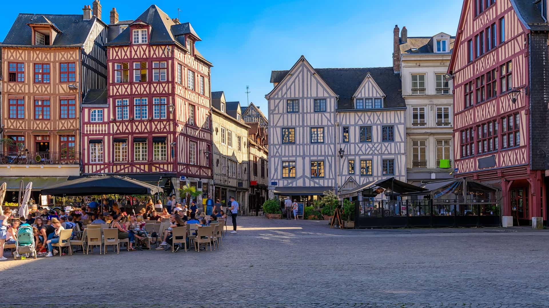 "A bustling summer day in Rouen, Normandy, with people dining at an outdoor cafe in front of historic, half-timbered houses in the Place du Vieux Marché. "