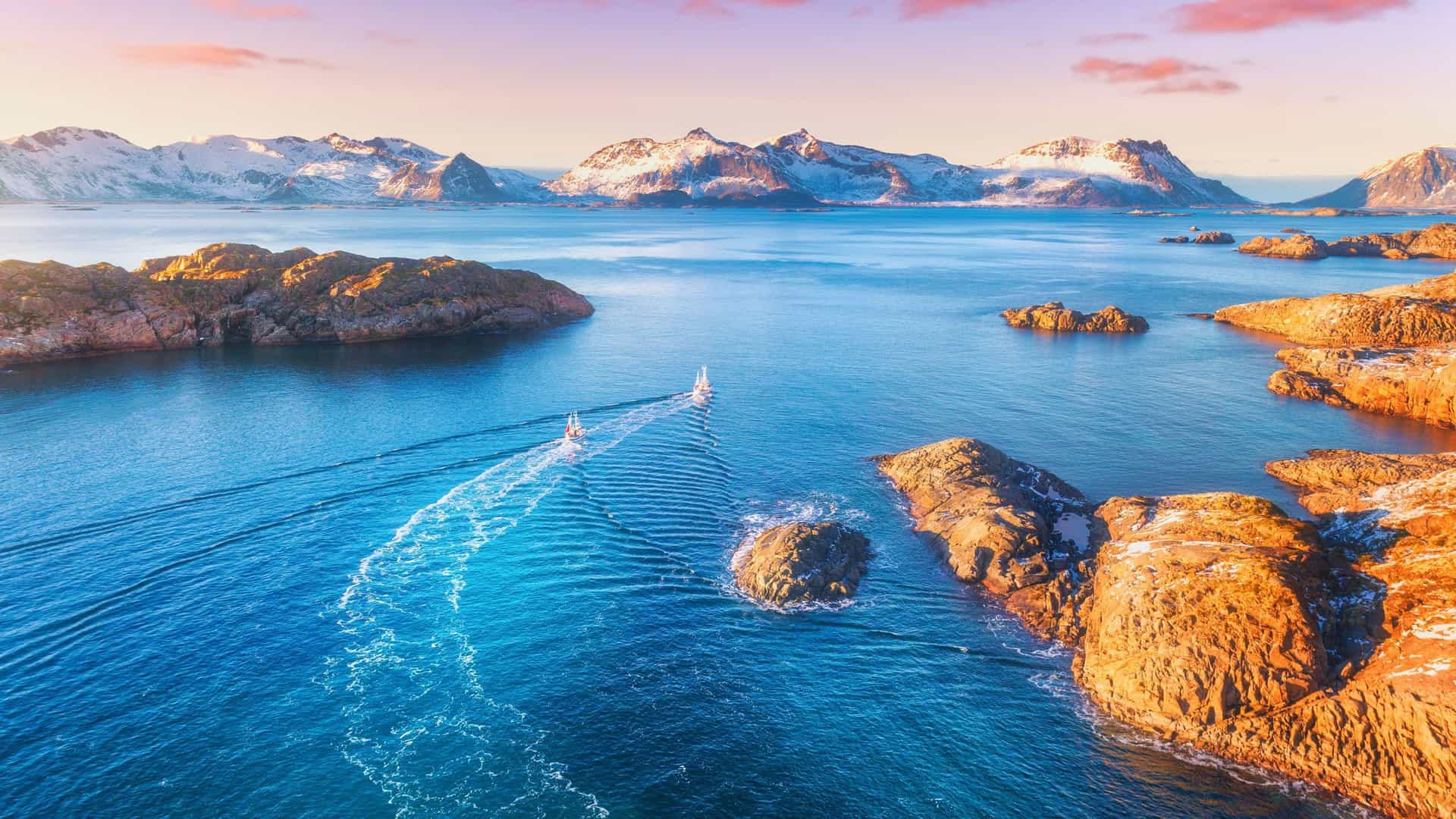 A scenic view of the Norwegian Sea with small sailboats navigating through rocky islands. In the background, majestic snow-capped mountains rise under a beautiful pastel sky at sunset.