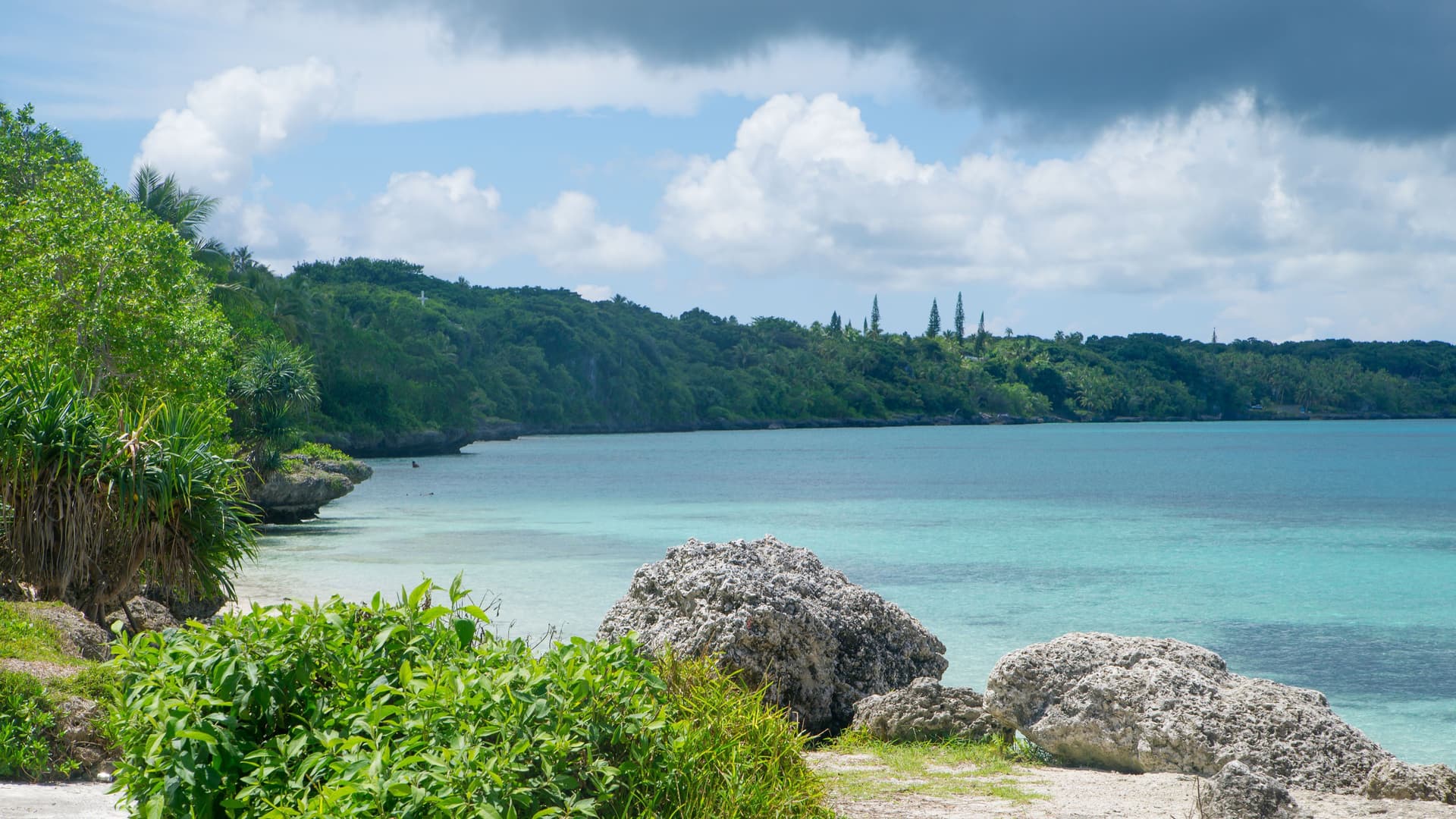 A stunning view of Anse Vata Bay in Noumea, New Caledonia, with crystal-clear turquoise water, lush green vegetation on the shoreline, and a partly cloudy sky overhead.