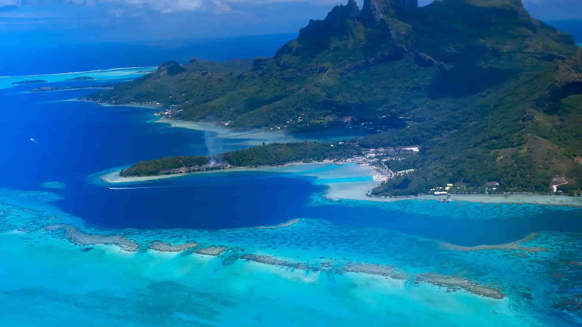 An aerial view of Nuku Hiva in the Marquesas Islands, French Polynesia, showing a volcanic island surrounded by a vibrant turquoise lagoon and coral reefs.