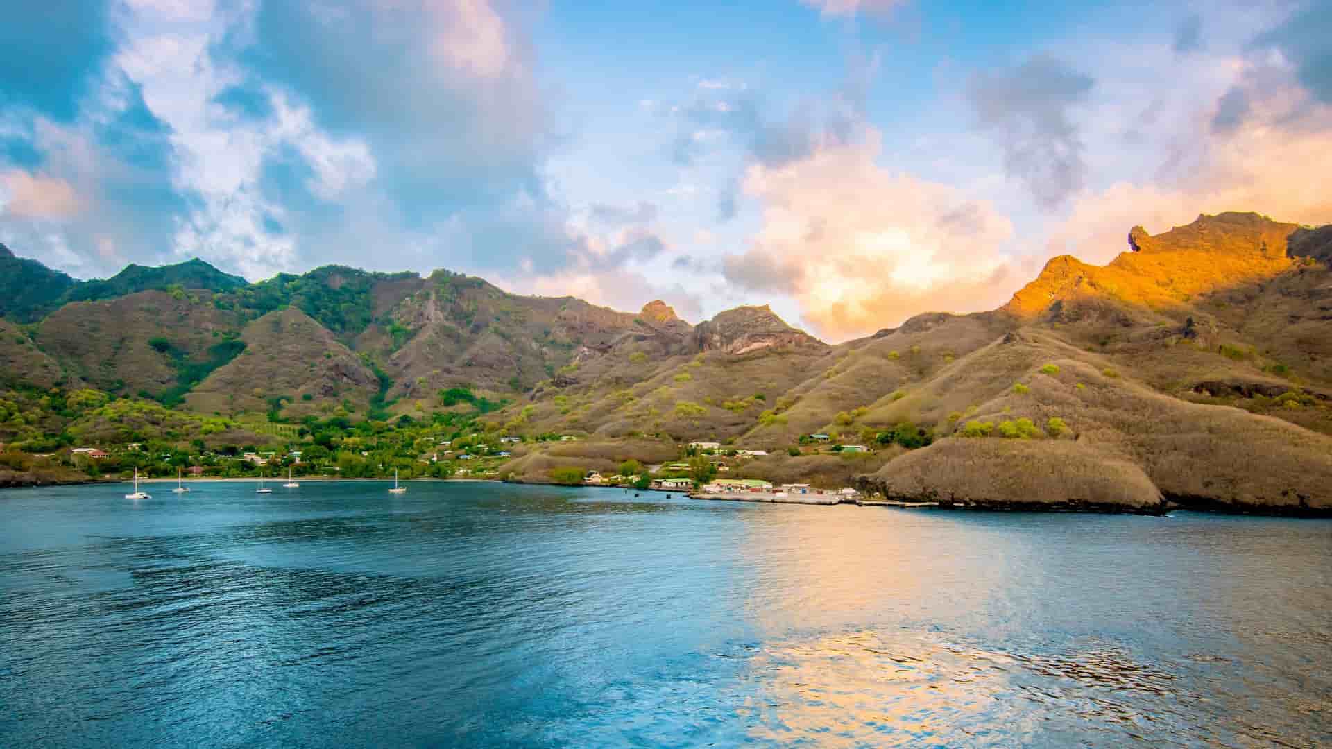 A panoramic view of Nuku Island, a volcanic Marquesas island, with steep, lush green hills and a small village lining a harbor with sailboats at sunset.