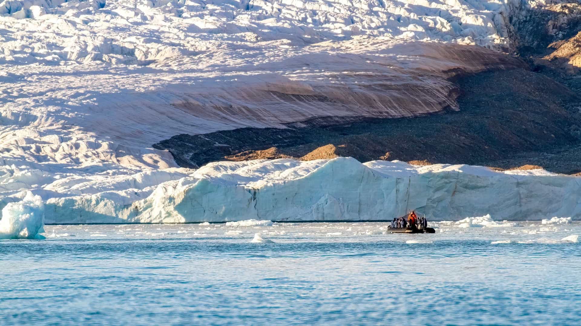 A group of tourists in a small black Zodiac boat cruising on the ice-filled waters of Croker Bay in Nunavut, with a massive glacier and ice cliffs in the background.