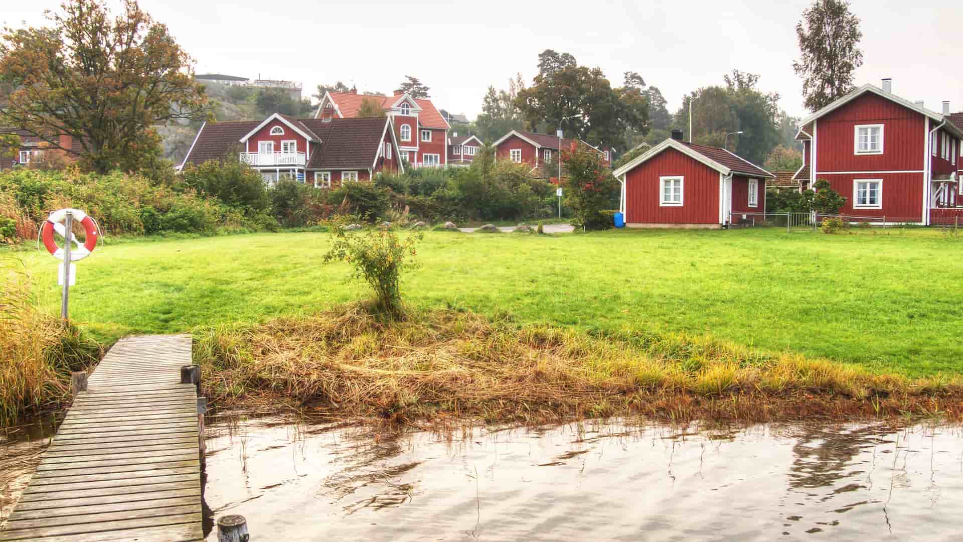 A charming scenic shot of a wooden dock leading to a lush green lawn with traditional red houses under a cloudy sky in Nynashamn, Sweden.