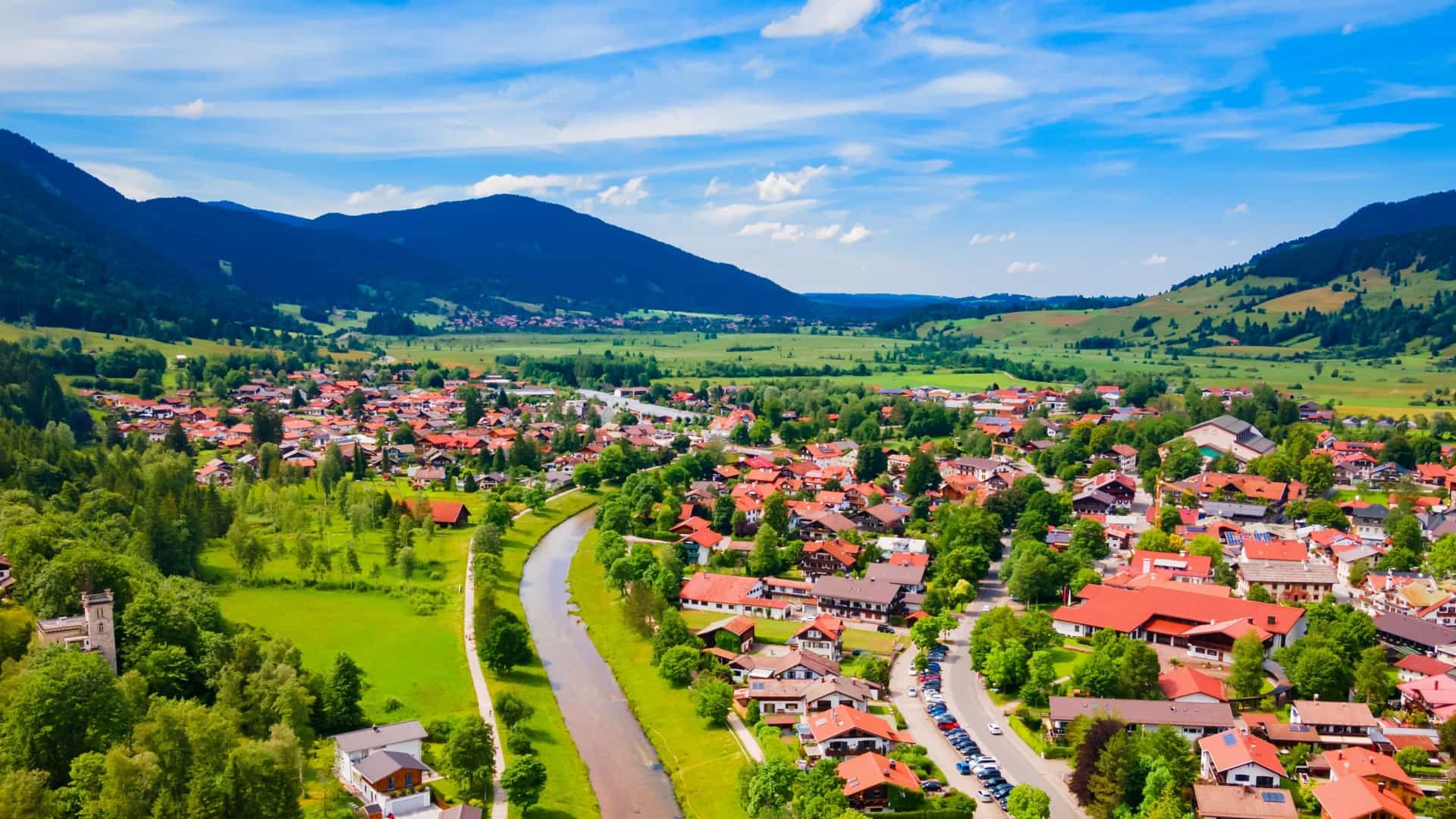 An aerial view of Oberammergau, a German village with red-roofed houses, situated in a green valley between rolling, forested mountains.