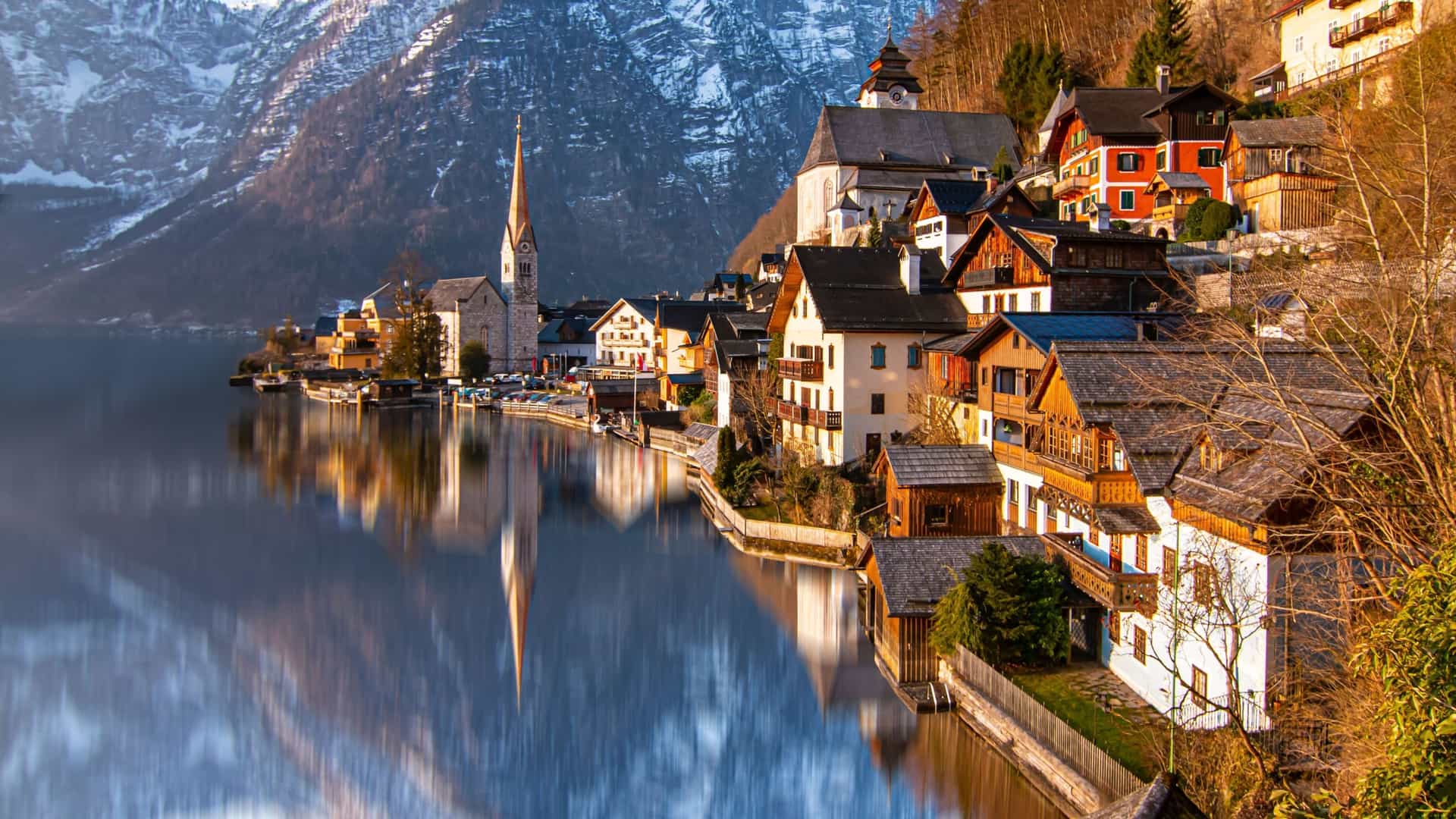 A scenic shot of the historic village of Hallstatt, Austria, nestled between a tranquil lake and snow-capped mountains, with its iconic church spire reflecting in the water.