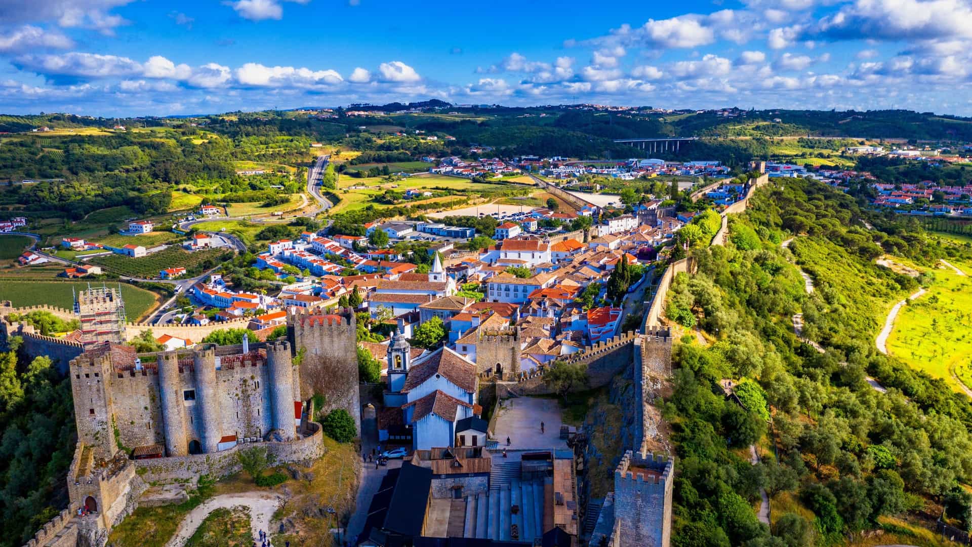 An aerial view shows the stone walls and medieval castle of Óbidos, Portugal, surrounding a cluster of white buildings with red roofs.