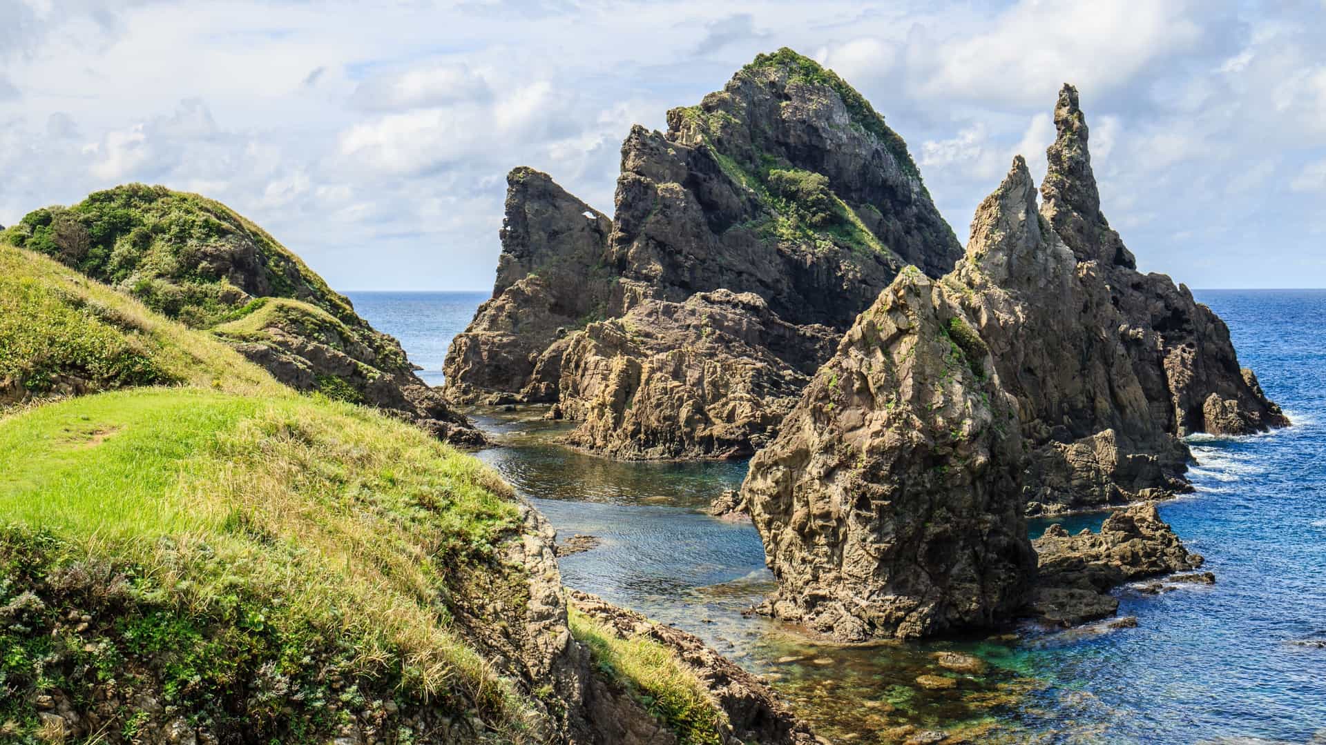 Jagged, dark volcanic rock formations and steep, grassy hills meet the clear blue ocean at the Oki Islands, Japan.