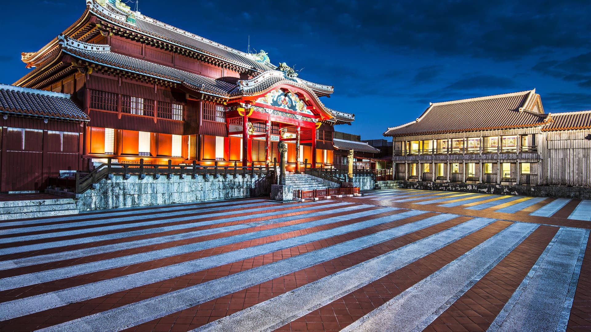 A night view of the illuminated Shuri Castle in Okinawa, Japan, with its traditional red and white striped courtyard, a major cultural landmark in the region.