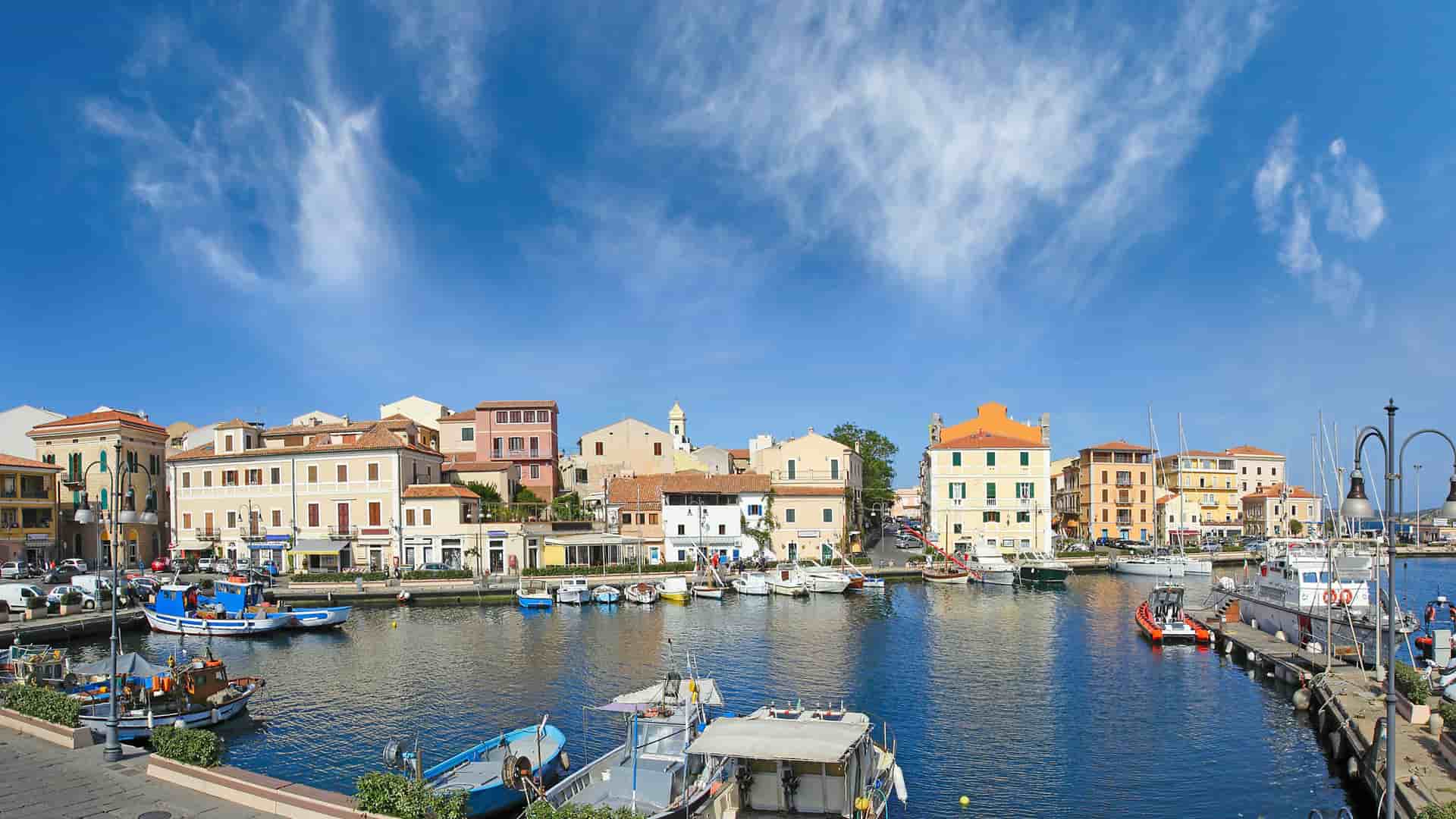 A vibrant panoramic view of the harbor in Olbia, Italy, with a wide variety of fishing boats and yachts docked in front of colorful waterfront buildings and a bright blue sky.