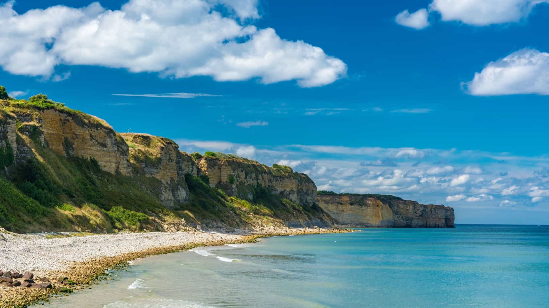 A panoramic shot of Omaha Beach in Normandy, France, showing the rugged cliffs and a pebble beach stretching along the tranquil coastline under a beautiful blue sky with white clouds.