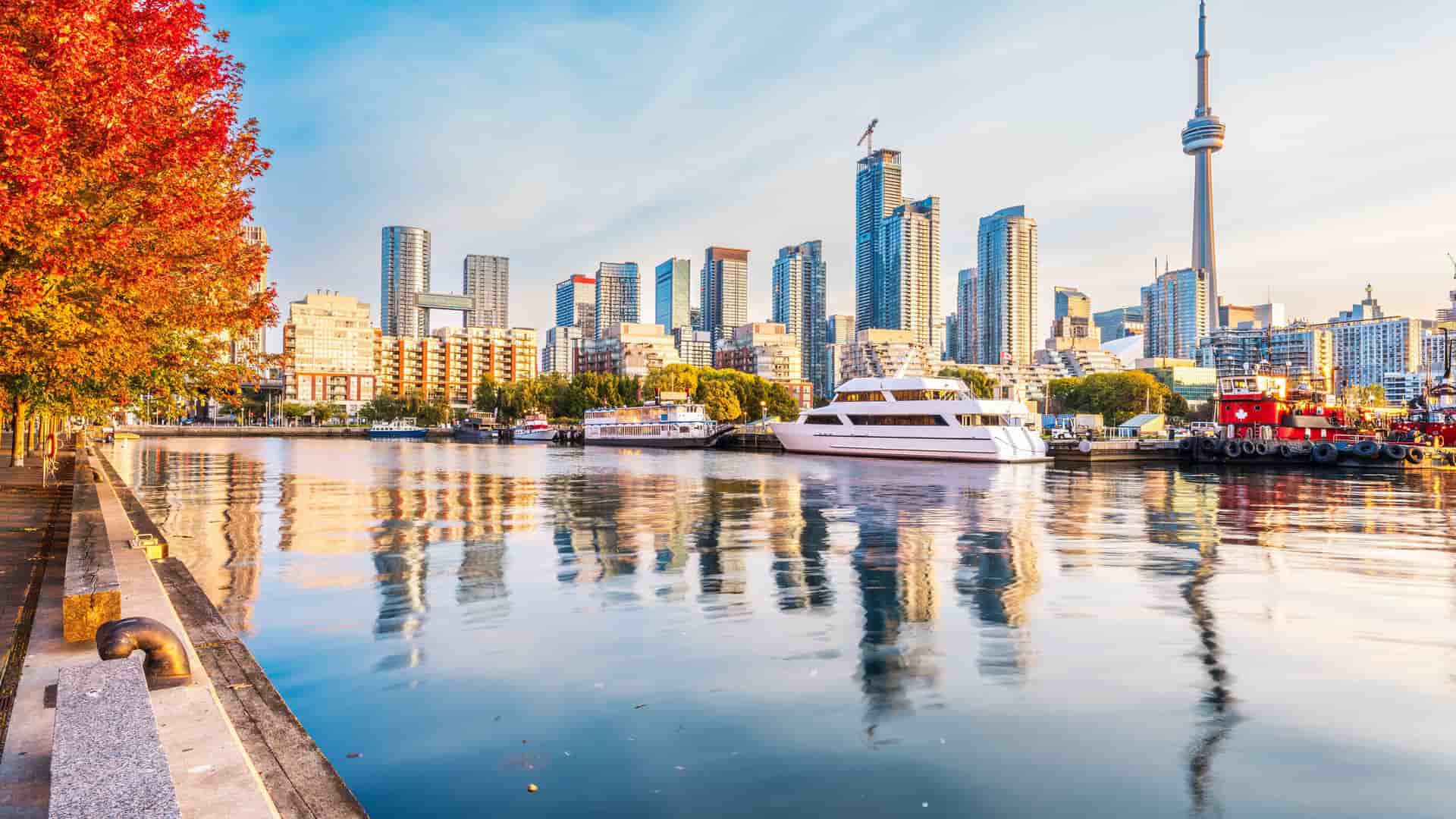 "A vibrant autumn panoramic view of the Toronto skyline at sunrise, showing the CN Tower, yachts, and the cityscape reflecting off the calm waters of Lake Ontario. "