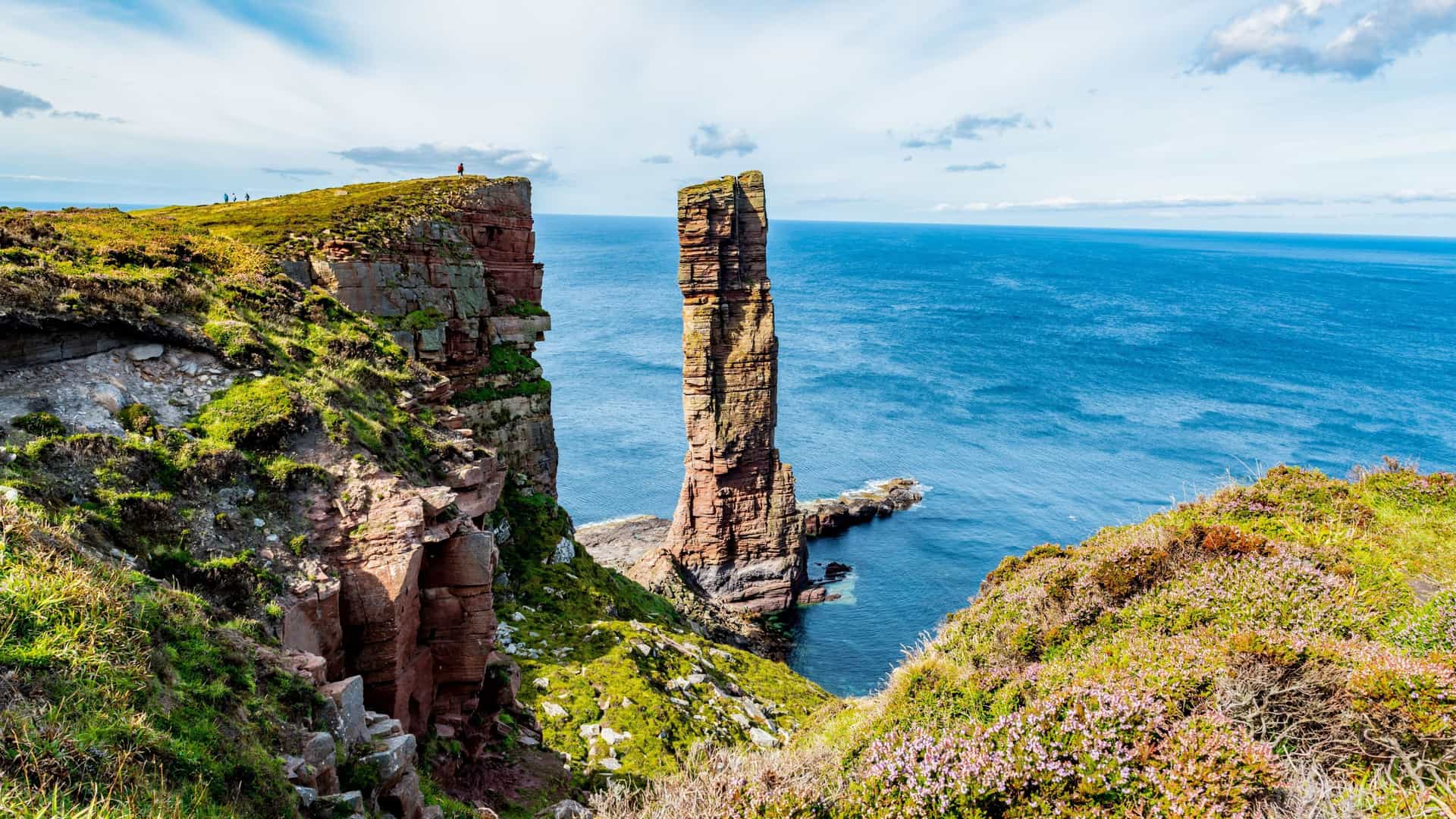 A stunning landscape shot of the Old Man of Hoy, a dramatic red sandstone sea stack on the coast of the Orkney Islands, with waves crashing against the base.