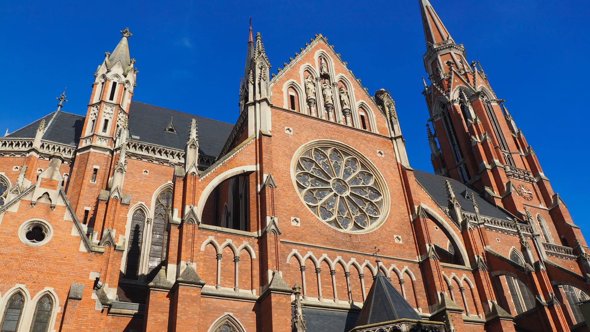 A striking low-angle view of the neo-Gothic Church of St. Peter and St. Paul in Osijek, Croatia, showcasing its intricate rose window, brick facade, and towering spires against a blue sky.