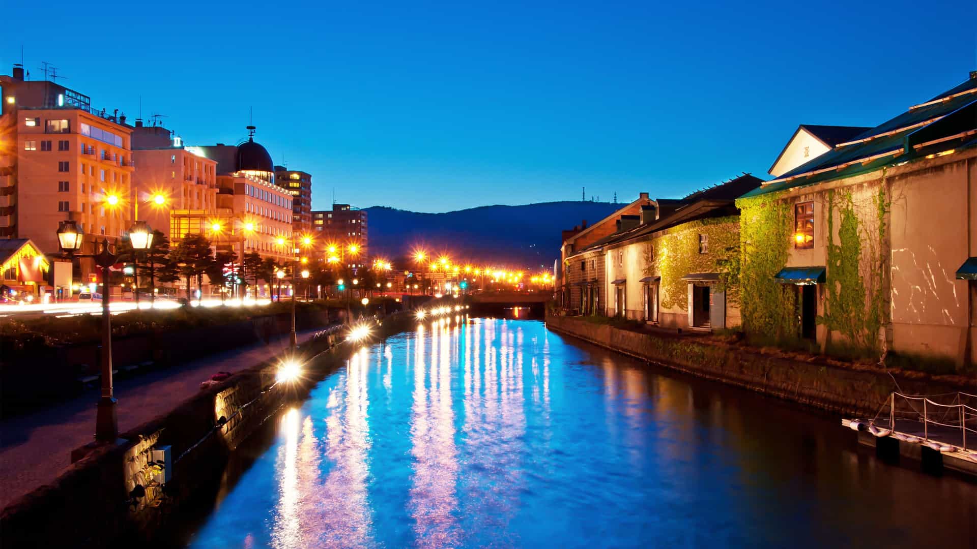 A stunning nighttime shot of the Otaru Canal in Japan, with historic warehouses and modern buildings illuminated on both sides, their lights reflecting beautifully on the water.