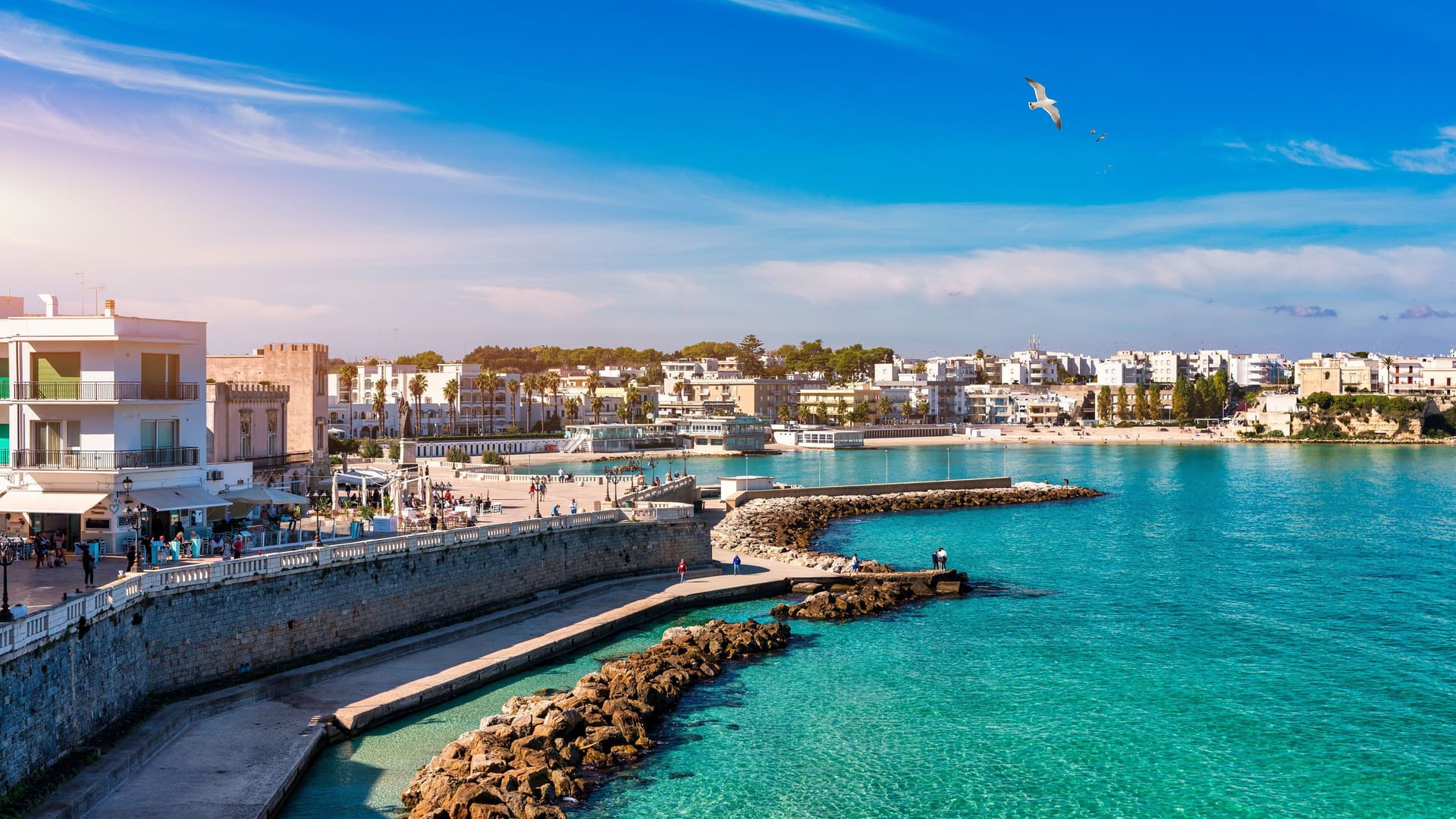 A picturesque view of Otranto, Italy, featuring a promenade along the turquoise-blue sea, with white buildings and a sandy beach under a bright blue sky.