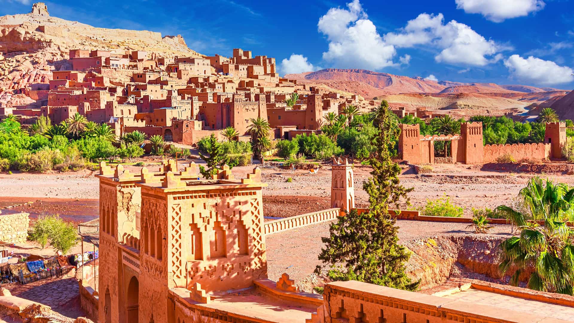 "A panoramic shot of the historic Aït Benhaddou, a fortified earthen city, or ksar, in Ouarzazate, Morocco, with palm trees and a lush green oasis at its base. "