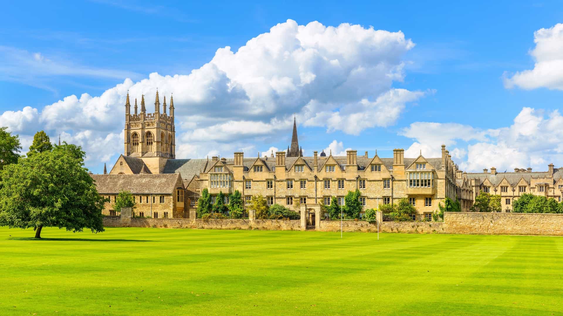 The Merton College complex, featuring medieval towers and classic limestone buildings, stands behind a wide, perfectly manicured green lawn under a cloudy blue sky.