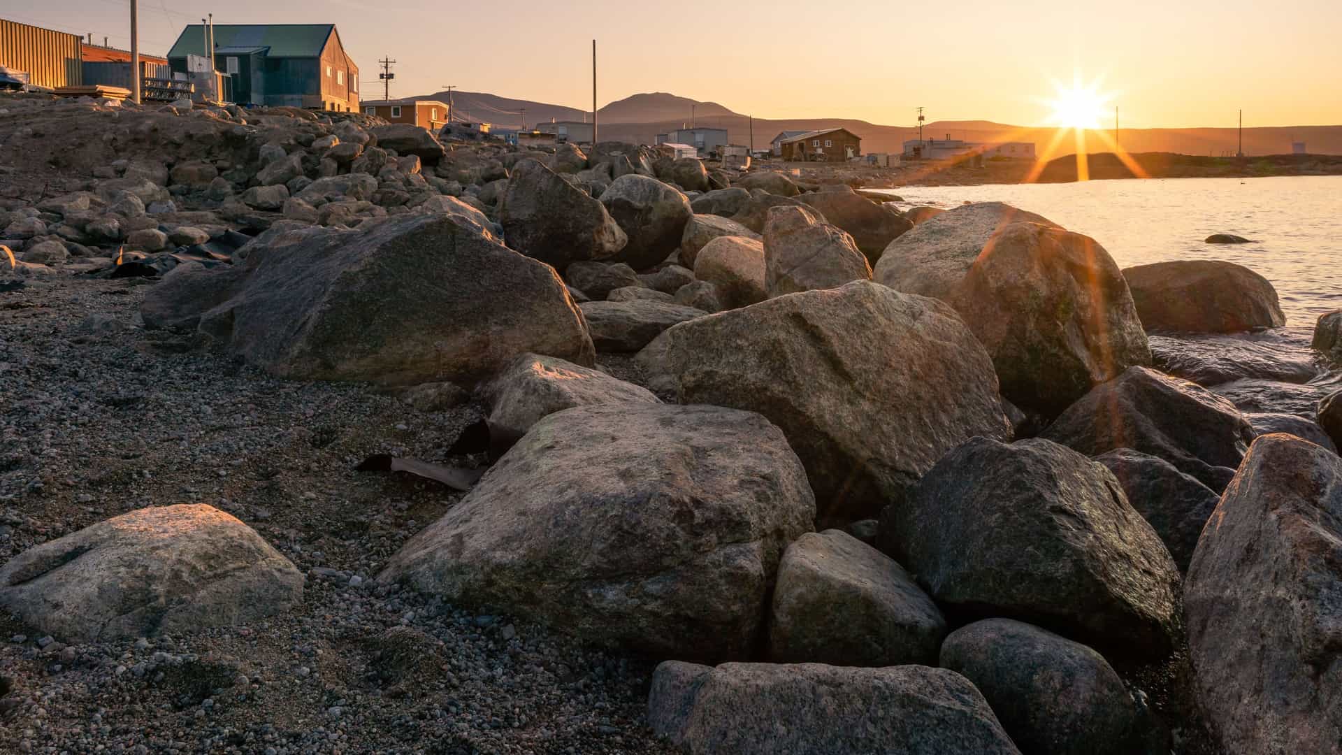 A close-up view of the rocky beach of Padloping Island, Nunavut, with a few houses and buildings in the background and the sun setting over the water.