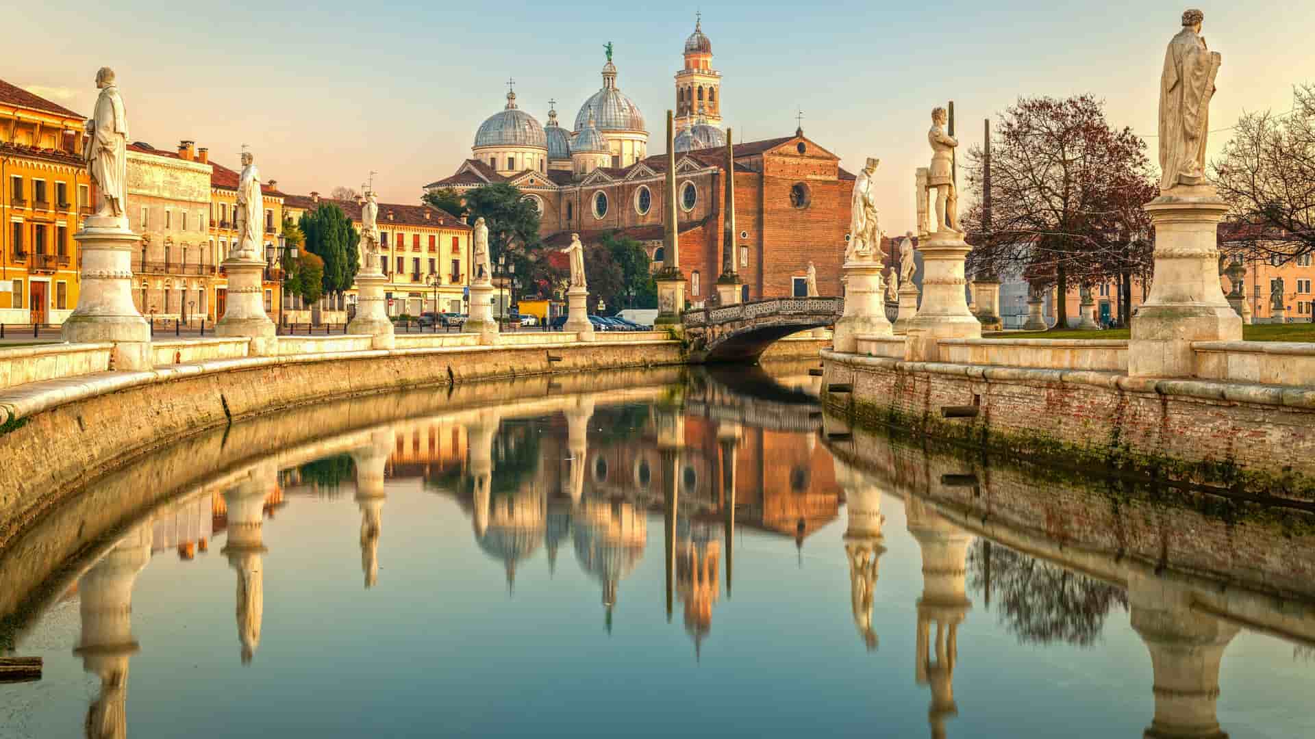 "A panoramic view of the historic Prato della Valle, one of Europe's largest squares, in Padua, Italy, with its central canal, statues, and the majestic Basilica of Santa Giustina reflected in the water. "