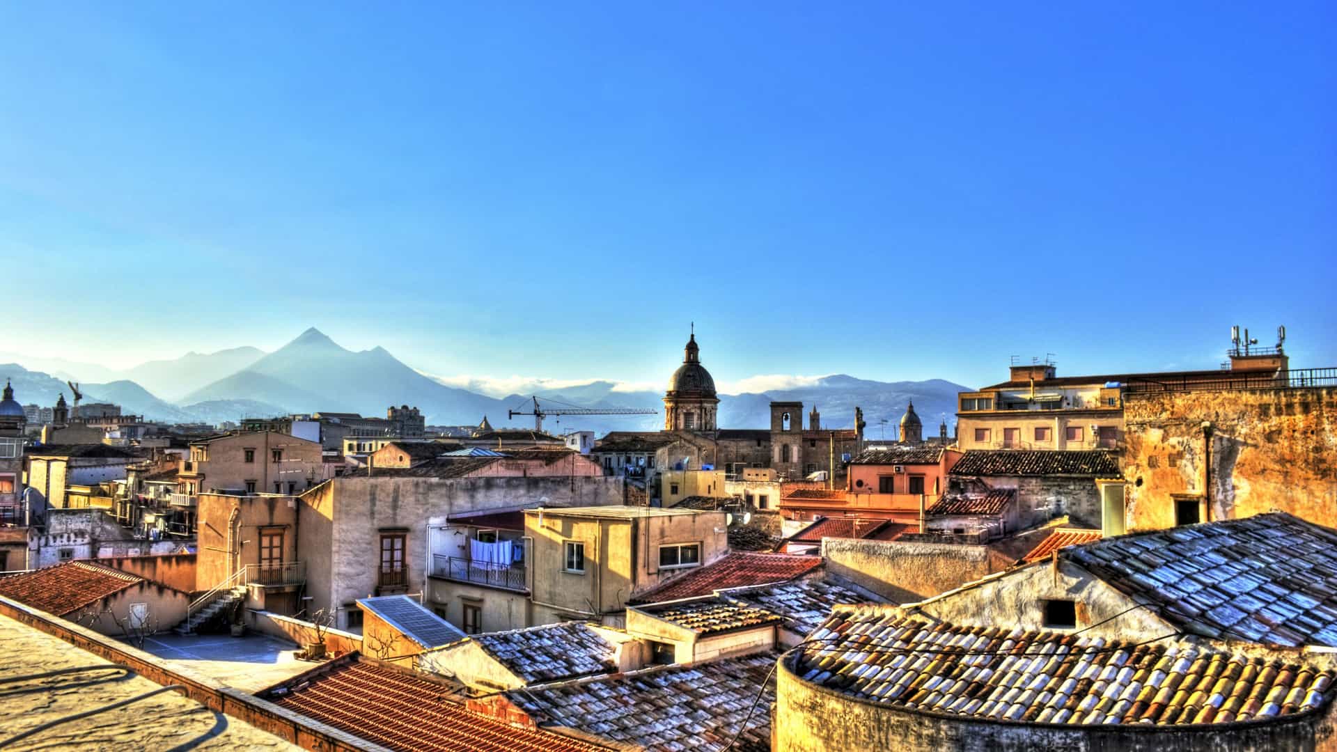 A high-angle view of the historic city of Palermo, Sicily, with a clear blue sky, buildings and rooftops in the foreground, and mountains in the distance.