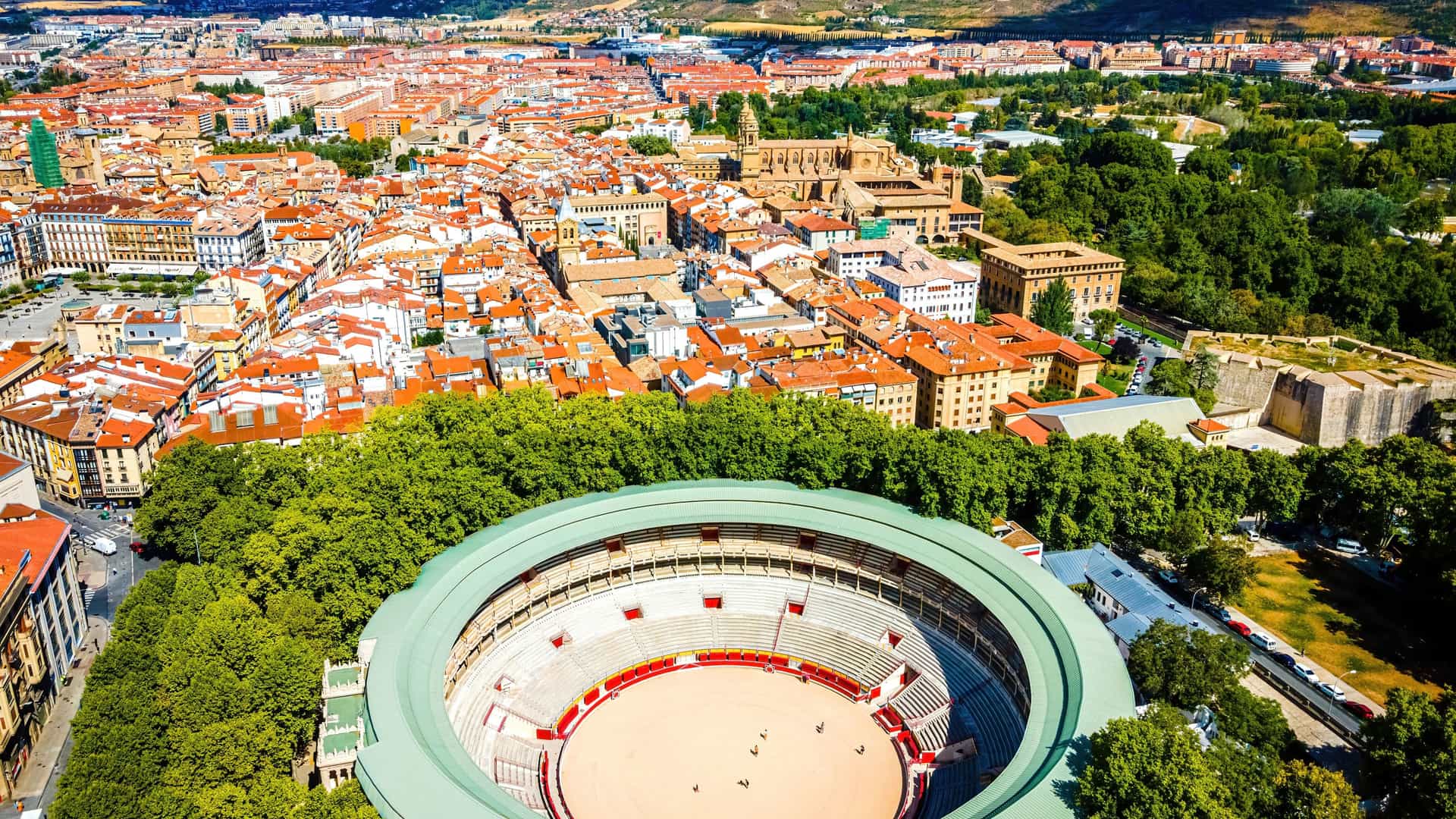 "An aerial panoramic shot of the historic Plaza de Toros, the bullring in Pamplona, Spain, with the city's old town and surrounding green trees and landscape in the background. "