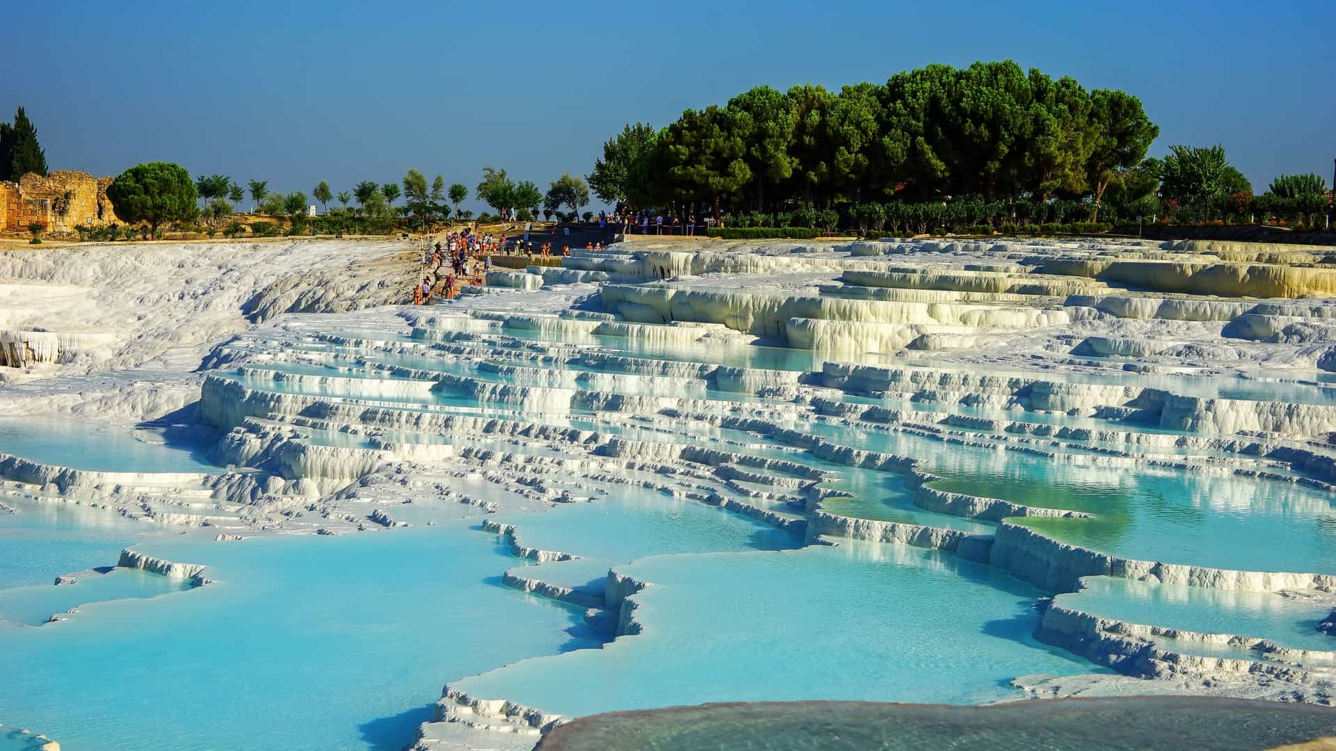 "A stunning wide-angle shot of the iconic Pamukkale thermal pools in Turkey, with people relaxing in the blue terraced waters and lush green trees in the background. "