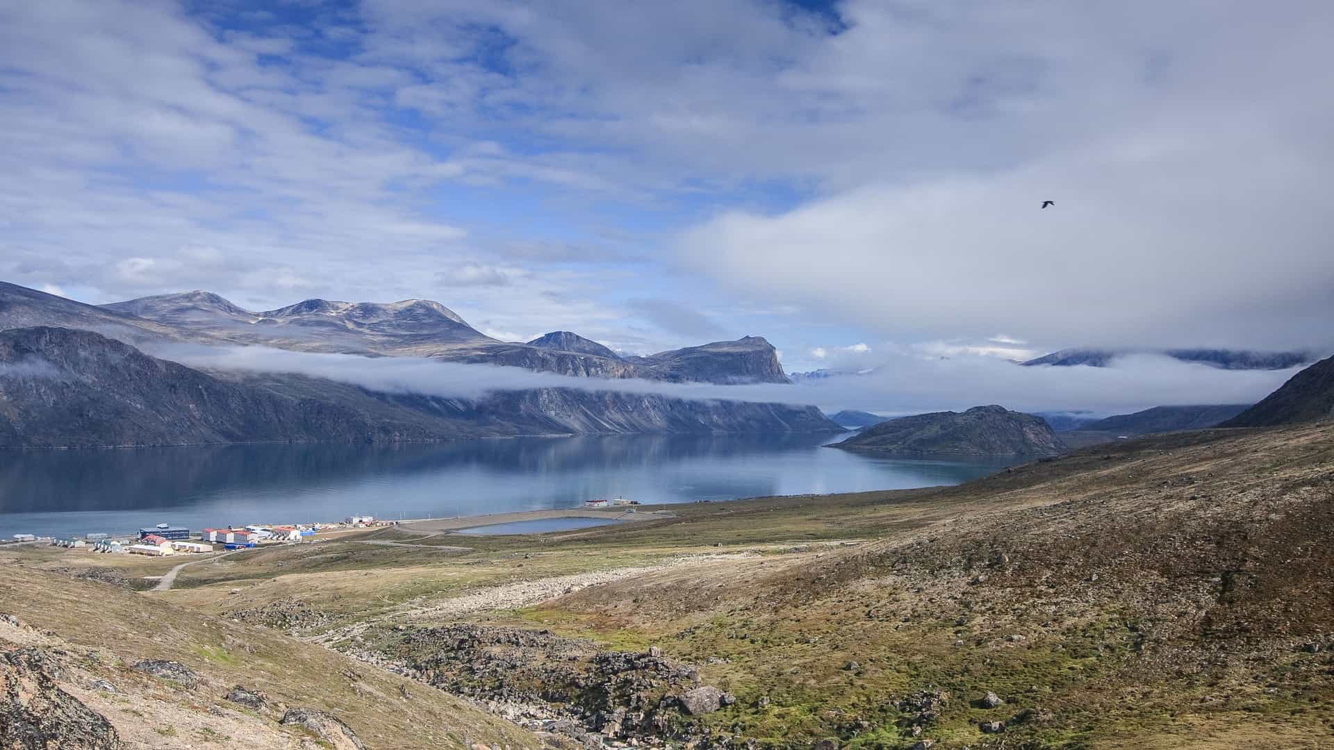 A beautiful scenic shot of the fjord in Pangnirtung, Nunavut, with small buildings along the shore, a huge lake, and clouds obscuring the tops of the mountain peaks.