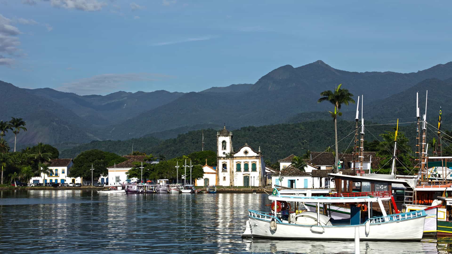 A view of the historic town of Paraty, Brazil, with boats docked in the harbor, colonial buildings, and mountains in the distance.