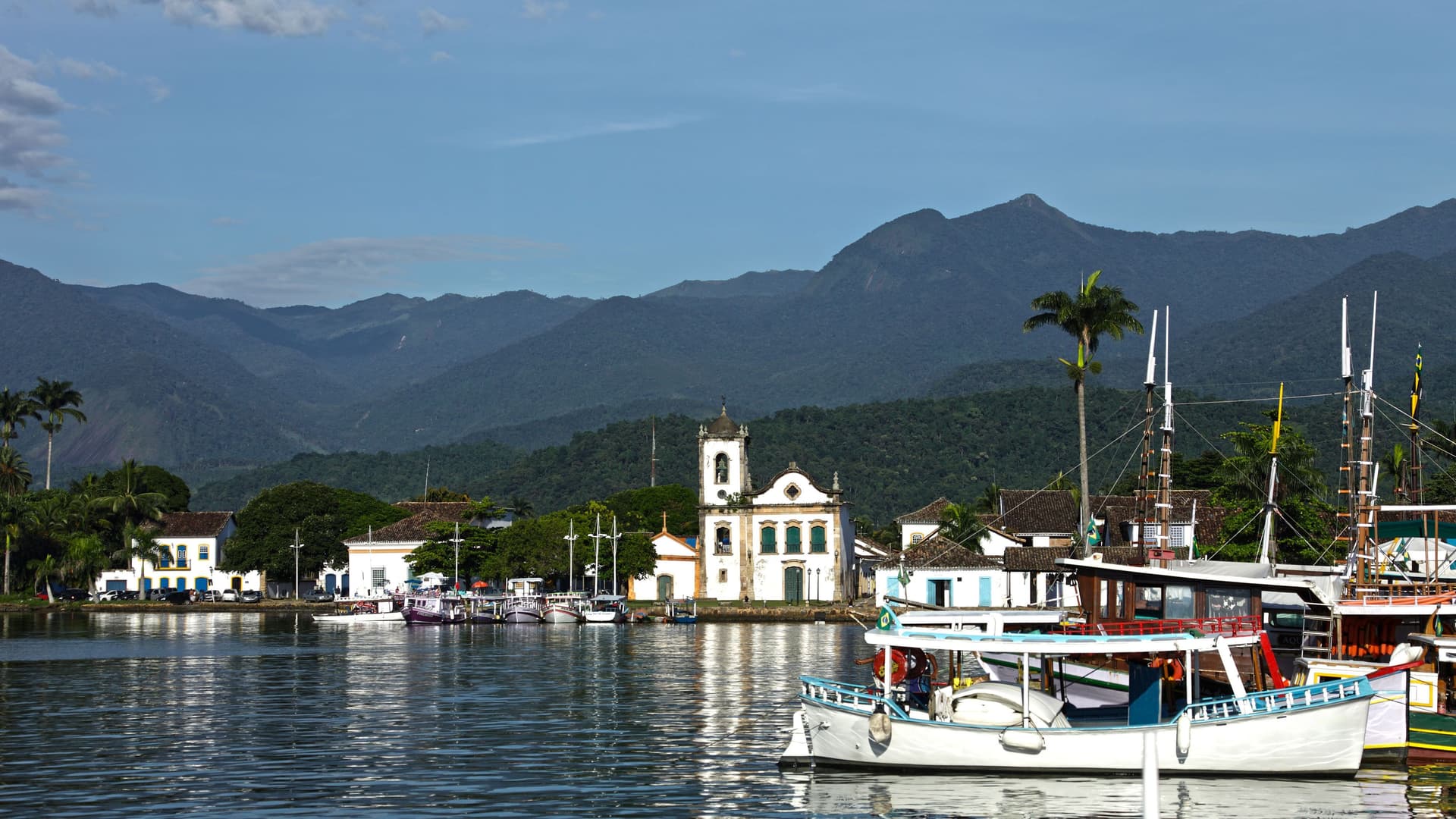 A view of the historic town of Paraty, Brazil, with boats docked in the harbor, colonial buildings, and mountains in the distance.