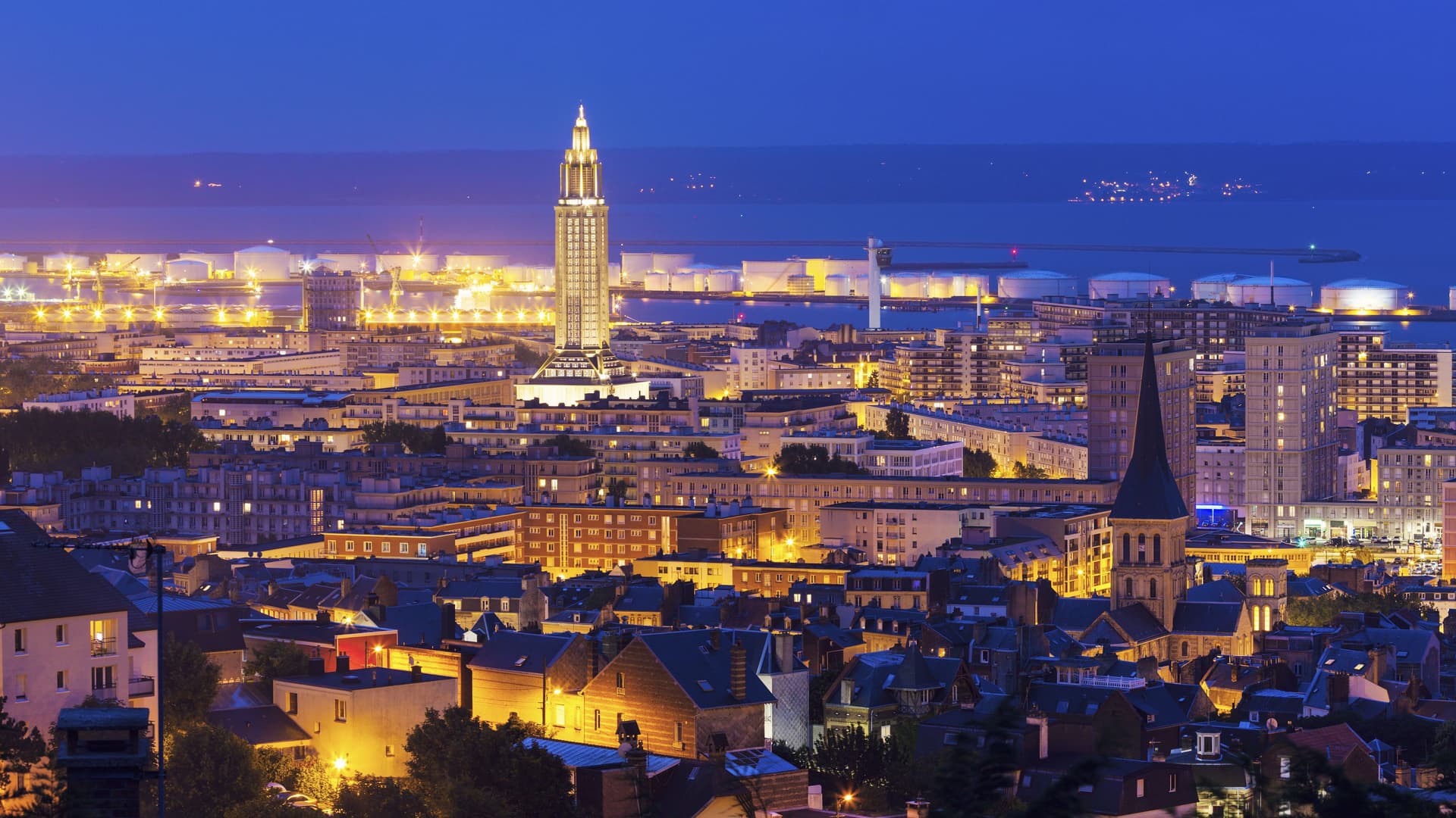 A wide-angle, nighttime view of Le Havre, France, with the tall Saint Joseph's Church steeple and the city lights illuminating the buildings and the harbor.