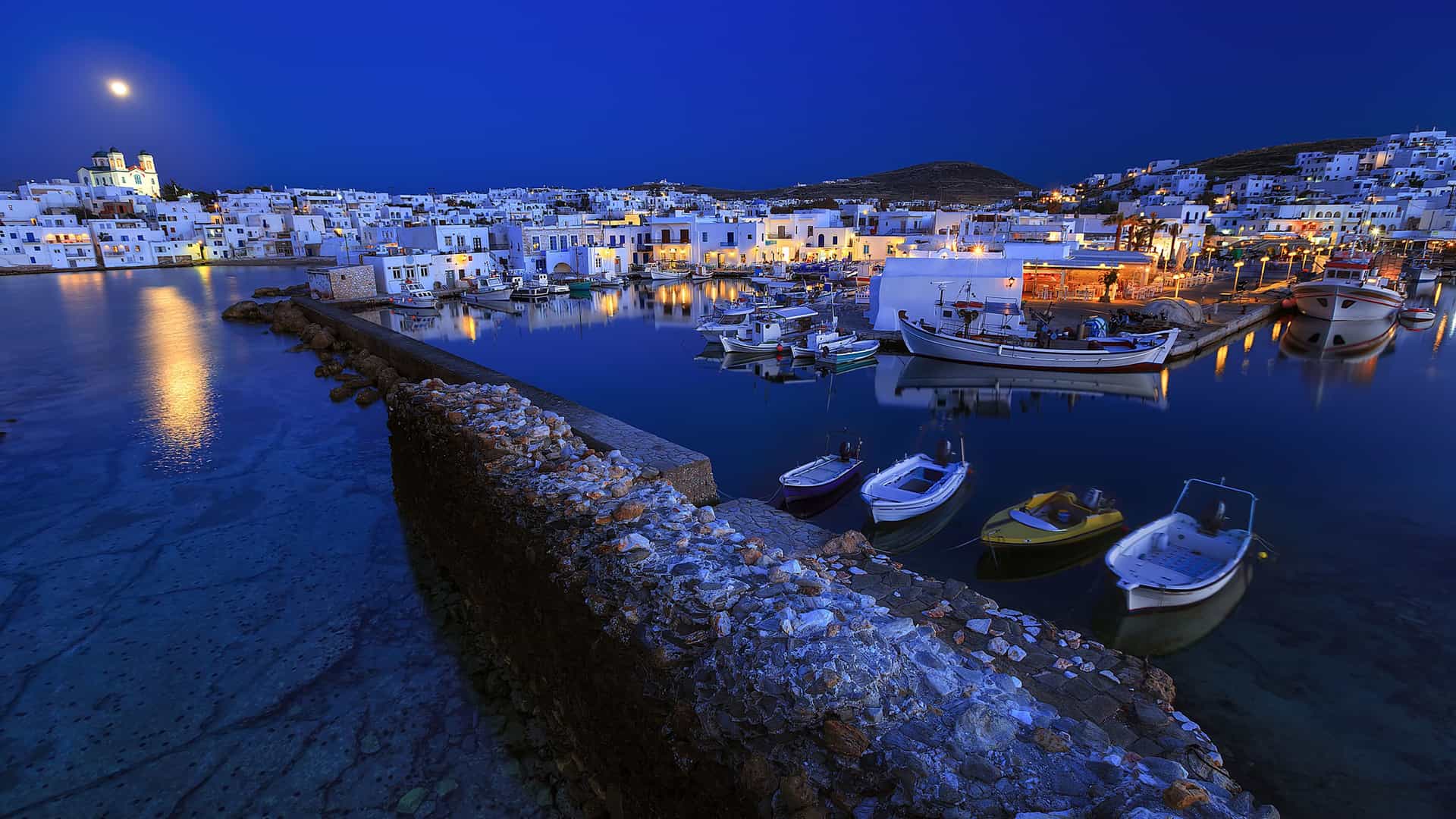 A nighttime shot of the Naoussa fishing harbor in Paros, Greece, with white buildings and boats docked in the calm water reflecting the lights and the moon.
