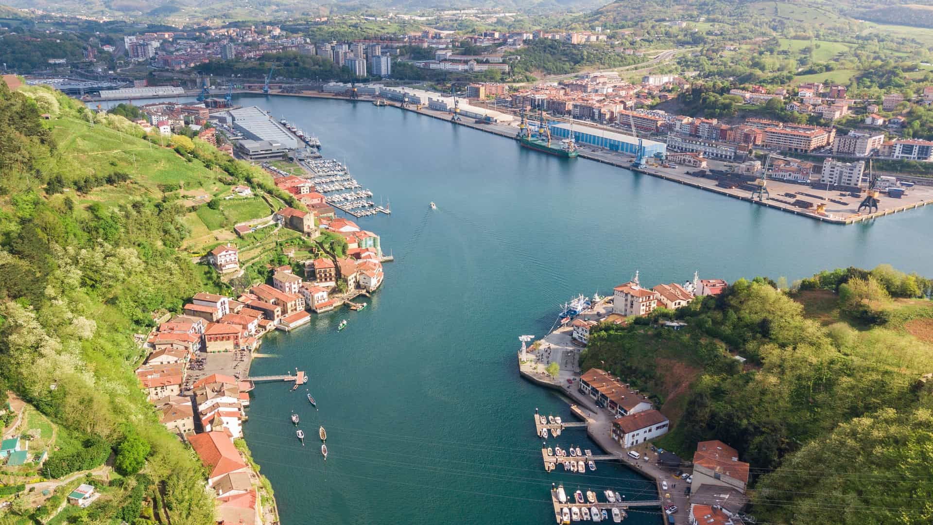 An aerial view of the Pasaia Bay (Bahía de Pasaia) inlet in the Basque Country, Spain. The image prominently features the narrow, winding entrance of the bay from the Cantabrian Sea, flanked by steep, green cliffs (Mount Ulia and Jaizkibel). Within the sheltered inlet, the dense, colorful cluster of houses of the two traditional fishing and port villages, Pasai Donibane (Pasajes San Juan) and Pasai San Pedro, is visible, situated directly along the waterfront. The bay transitions into the larger, industrialized port area further inland.