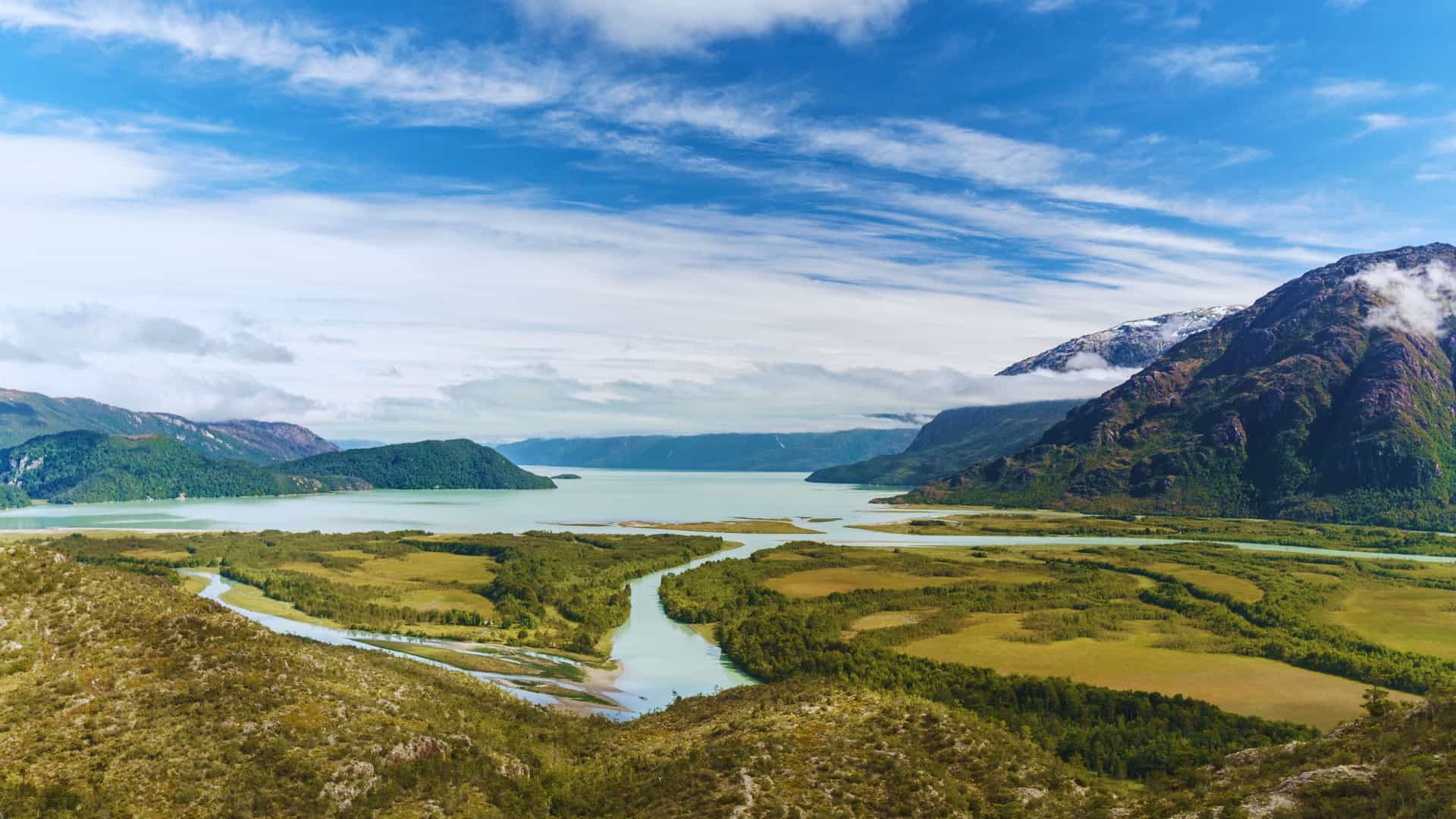 A panoramic view of the Patagonian fjords near the remote village of Caleta Tortel, Southern Chile. The dark, calm water of the fjord reflects the steep, rugged mountains and dense green forests that rise dramatically from the shore. The unique cypress wood stilt houses and boardwalks of the isolated village are visible along the coastline.