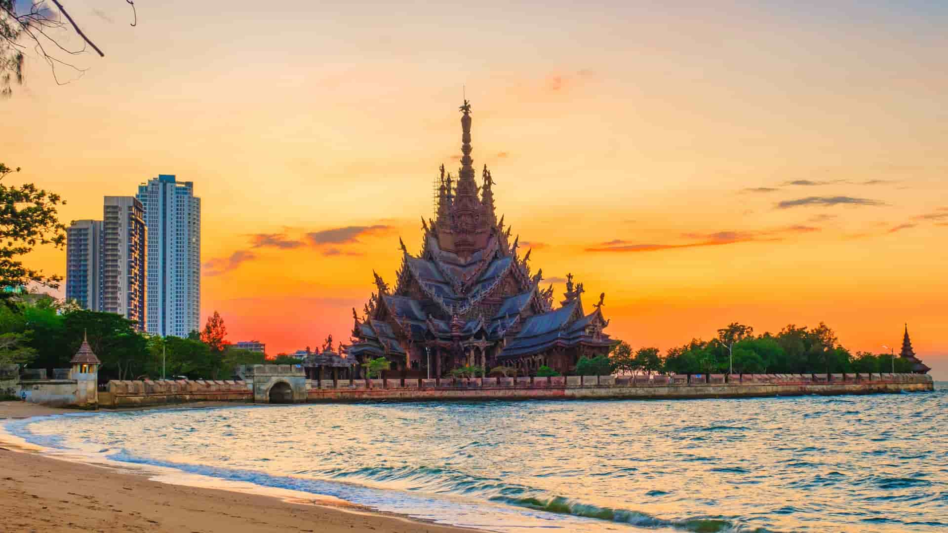 A striking coastal view in Pattaya featuring the ornate wooden Sanctuary of Truth silhouetted against a vivid orange‑pink sunset, with the beach and ocean in the foreground and modern high‑rise buildings rising in the distant background.