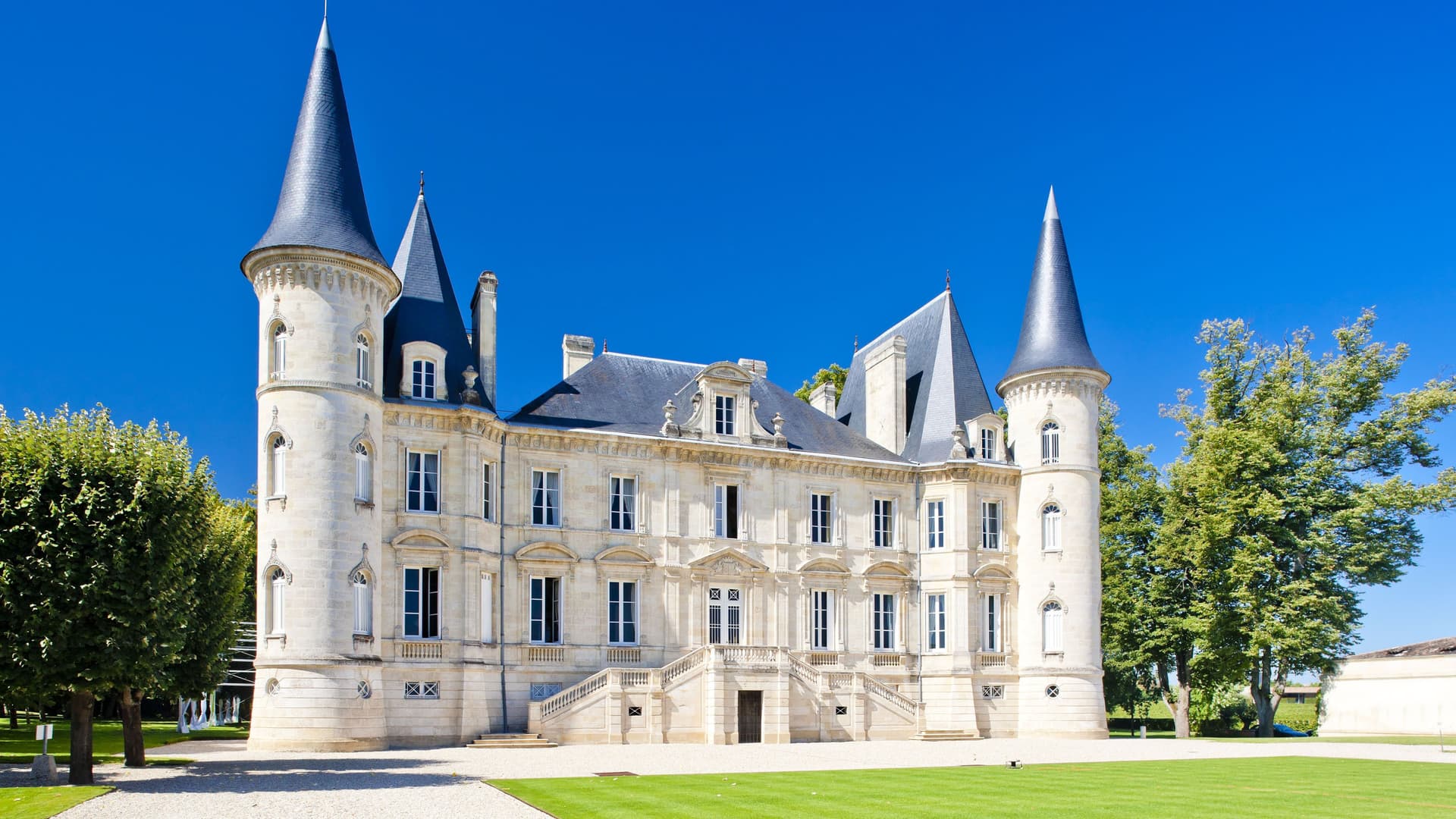 A grand, historic château with multiple turrets and a perfectly manicured lawn, the famous Château Pichon Baron, a beautiful landmark in Pauillac, France.