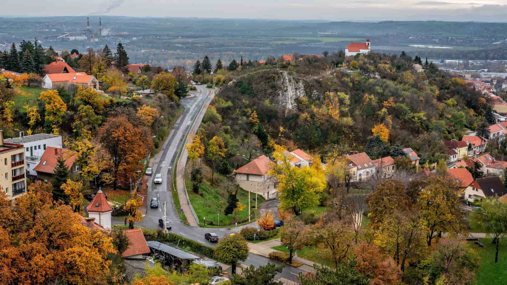 A scenic, elevated view of the city of Pécs, Hungary, with a winding road leading through a hillside neighborhood with red-roofed buildings and colorful autumn trees.