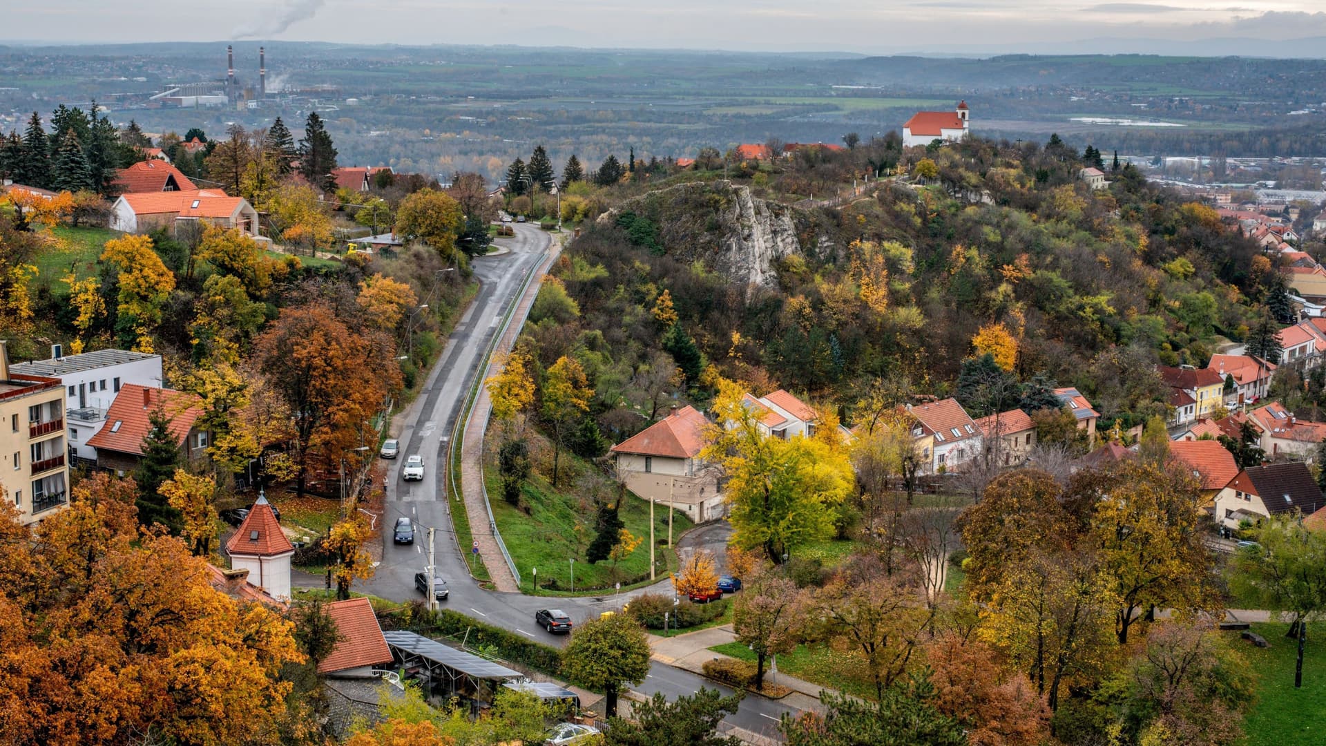 A scenic, elevated view of the city of Pécs, Hungary, with a winding road leading through a hillside neighborhood with red-roofed buildings and colorful autumn trees.