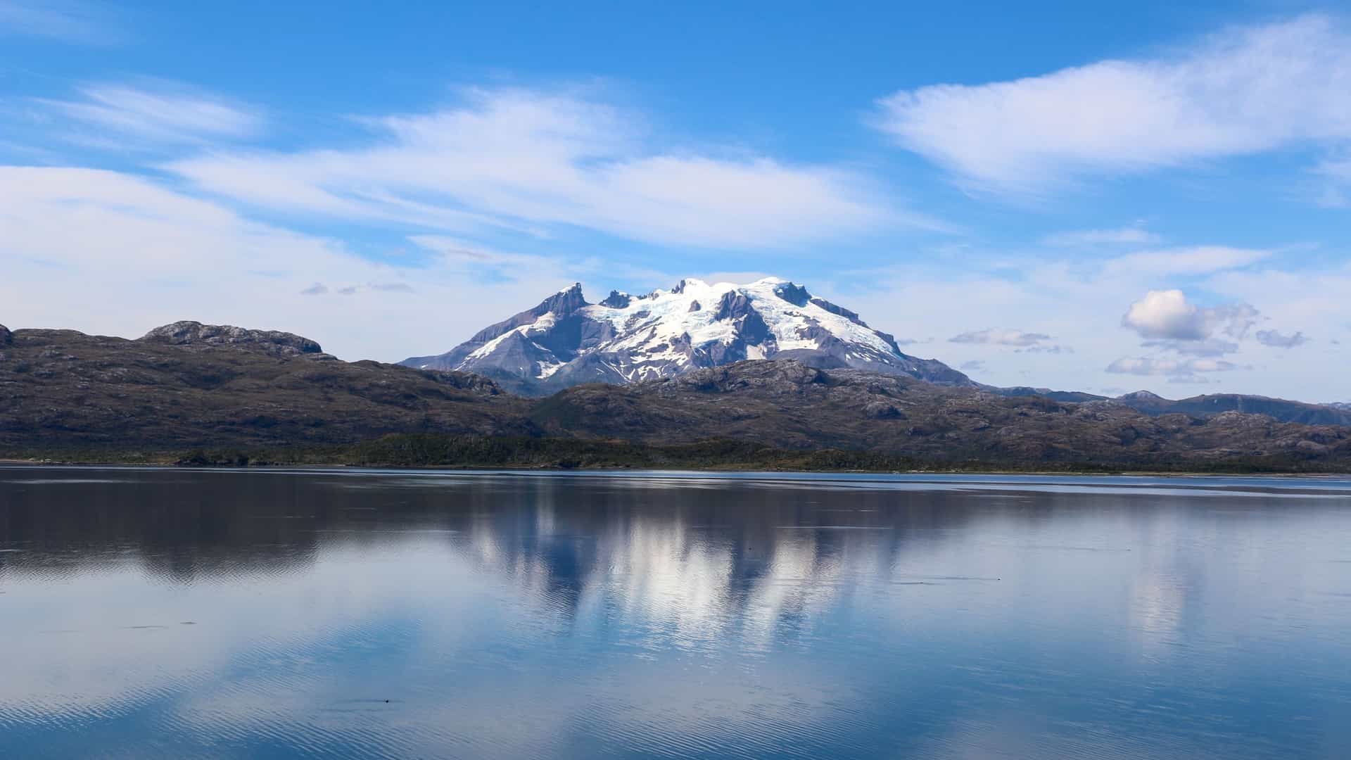 A scenic view of Peel Fjord in Chile, with a snow-capped mountain reflecting on the calm water and a cloudy blue sky in the background.