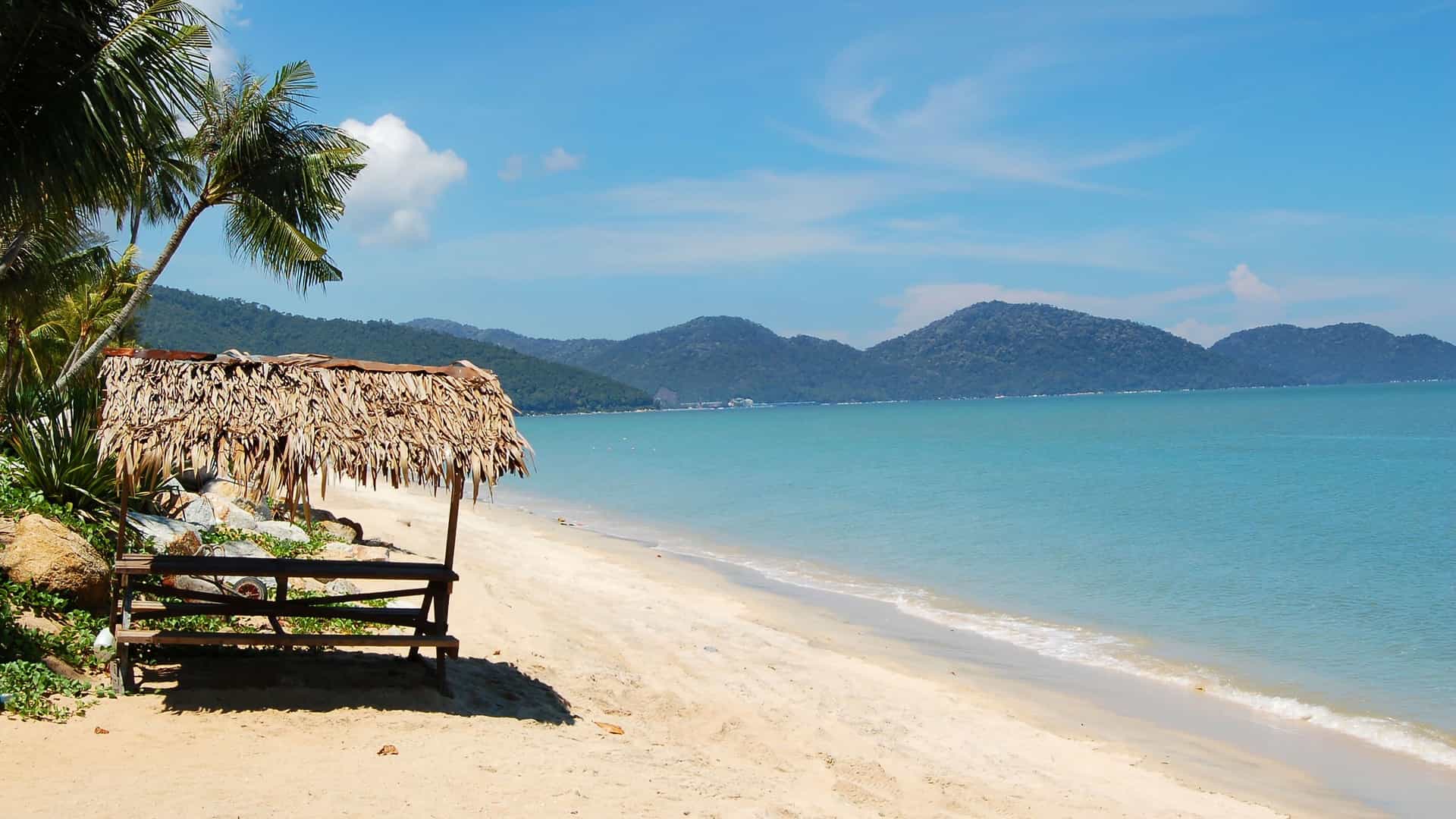 A view of the sandy beaches of Penang, Malaysia, with a thatched-roof hut on the shore, palm trees, and the clear blue ocean.