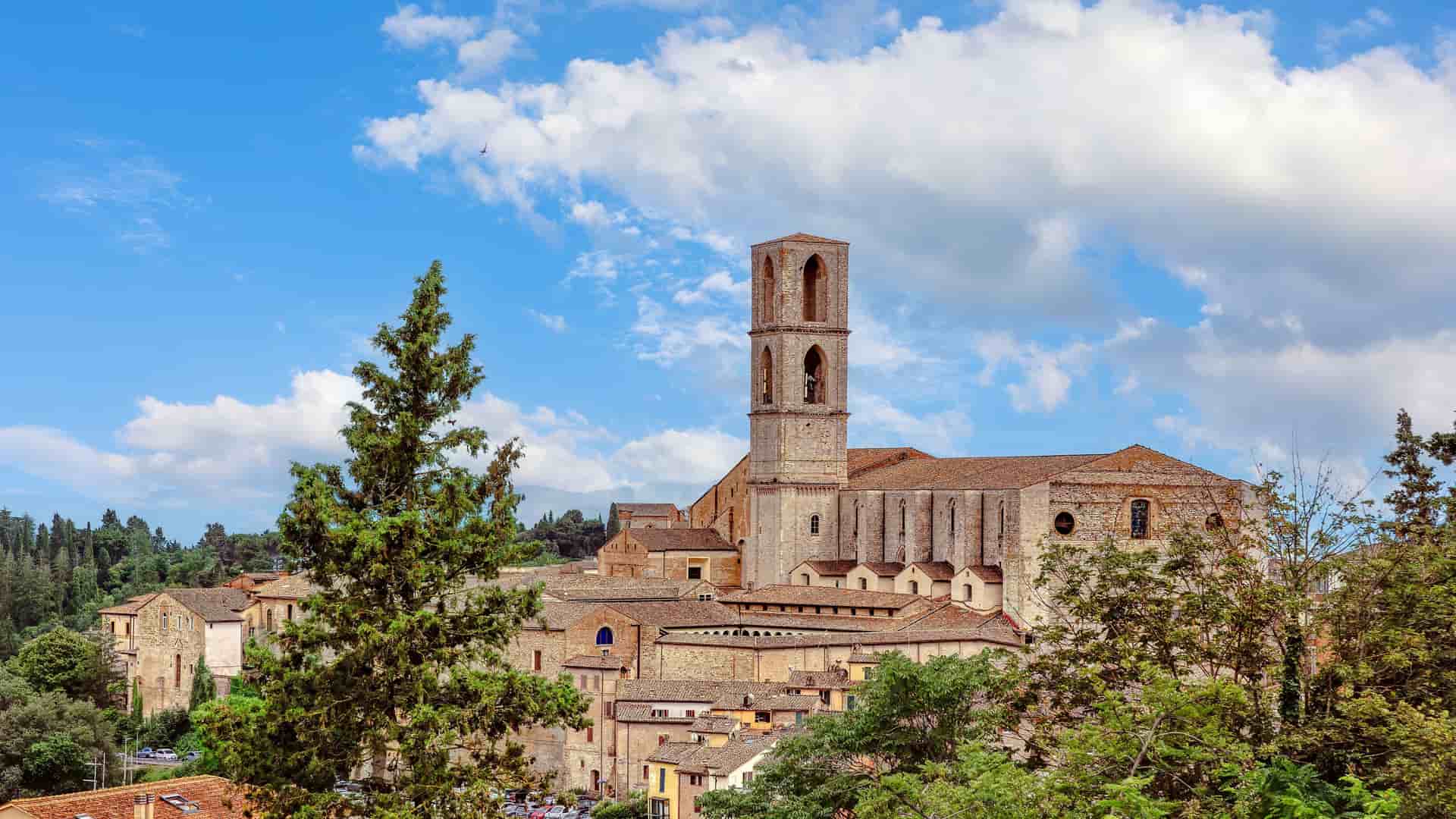 "A stunning wide-angle shot of the historic Abbey of San Pietro, a major landmark in Perugia, Italy, with its towering bell tower and medieval stone buildings surrounded by lush trees under a cloudy blue sky. "