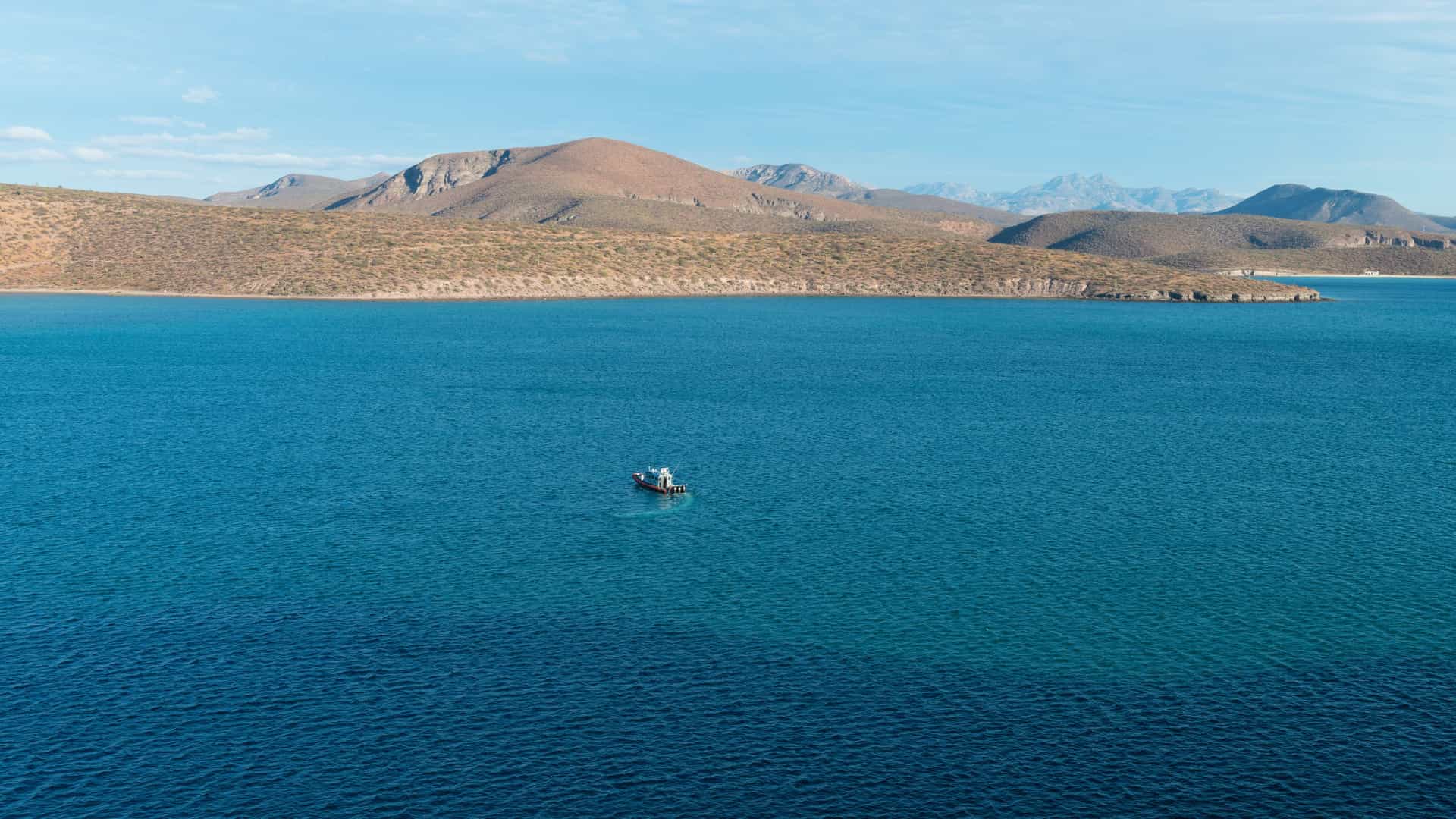 A calm coastal scene in Pichilingue with a lone small boat floating on deep blue water, rugged brown hills lining the distant shore, and soft clouds drifting across a bright sky.