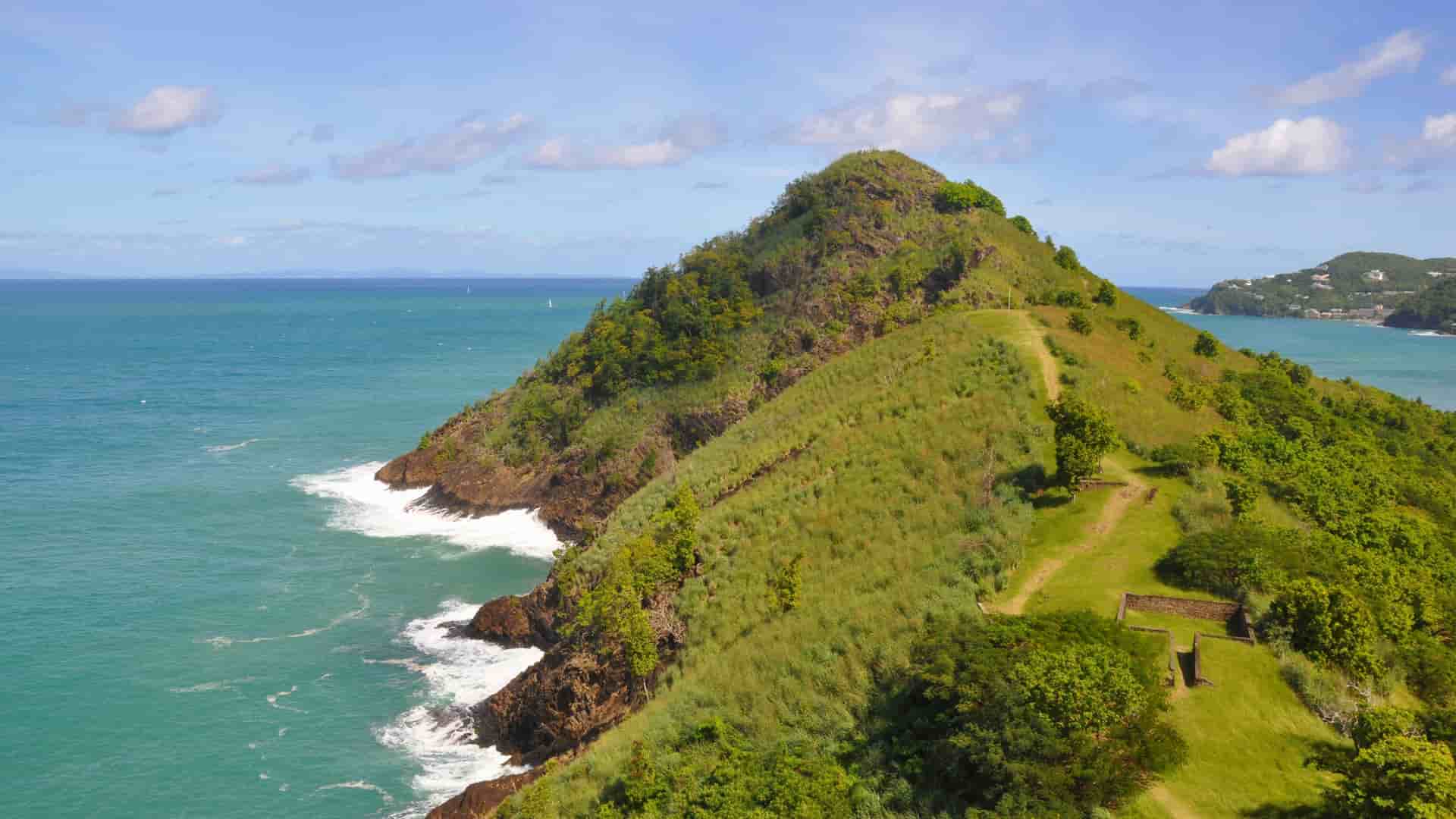 A view of Pigeon Island National Landmark, St. Lucia, showing a lush, green peninsula jutting out into the ocean with waves crashing on the rocks.