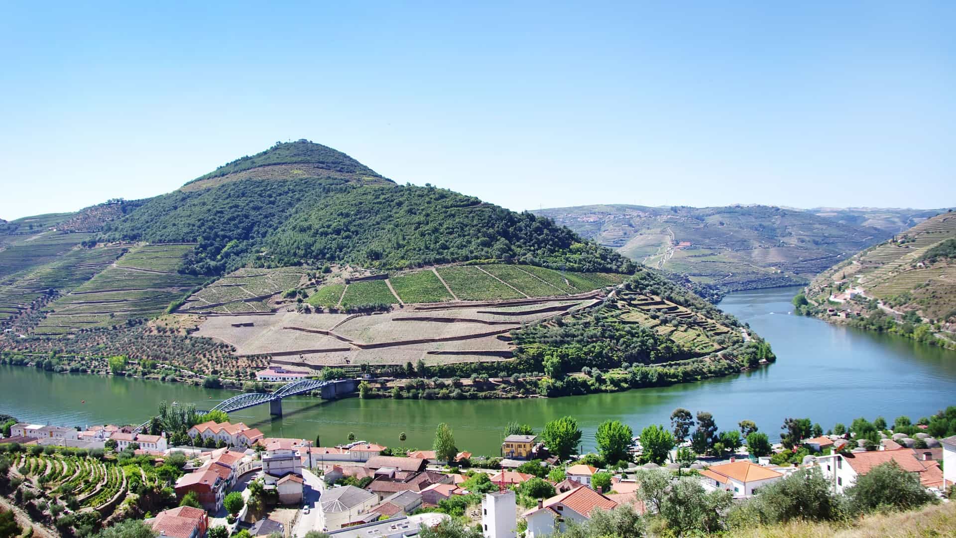 A panoramic view of the Douro River in Portugal, showcasing the terraced vineyards on the hillsides surrounding the small town of Pinhão, with a bridge crossing the water.